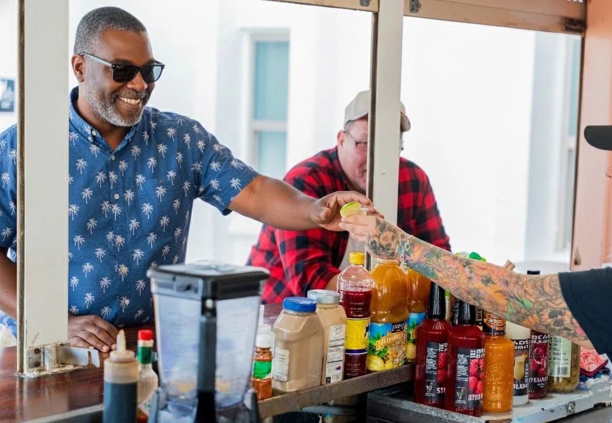 A man with sunglasses and a blue Hawaiian shirt is smiling while handing money through a window to a tattooed person with a call cap, in front of a counter with various condiments.