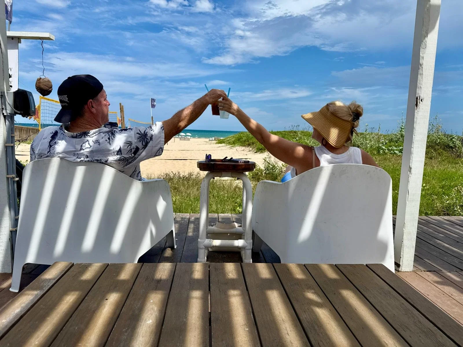 A man and woman sitting outside at a beachside restaurant or bar, clinking their drinks together in a toast, with the ocean and sky in the background.
