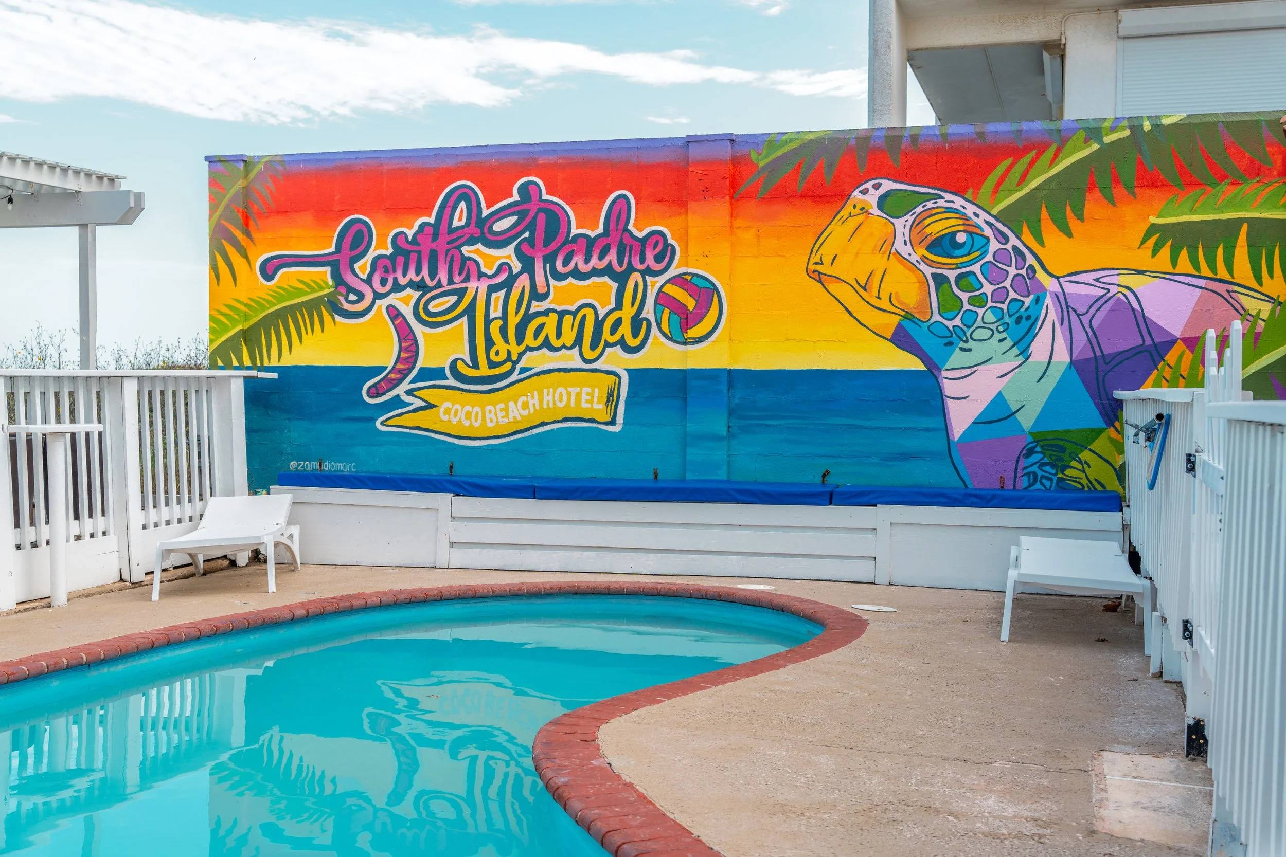 A colorful mural on a wall featuring the text 'South Padre Island' and 'Coco Beach Hotel', with a turtle and palm leaves, next to a swimming pool at sunset.