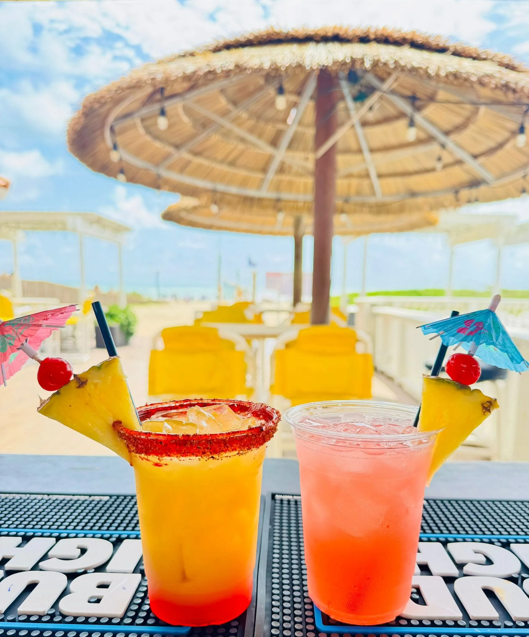 Two colorful tropical cocktails with pineapple slices, cherries, and umbrellas on a table at an outdoor beach bar with yellow chairs and a thatched umbrella under a partly cloudy sky.