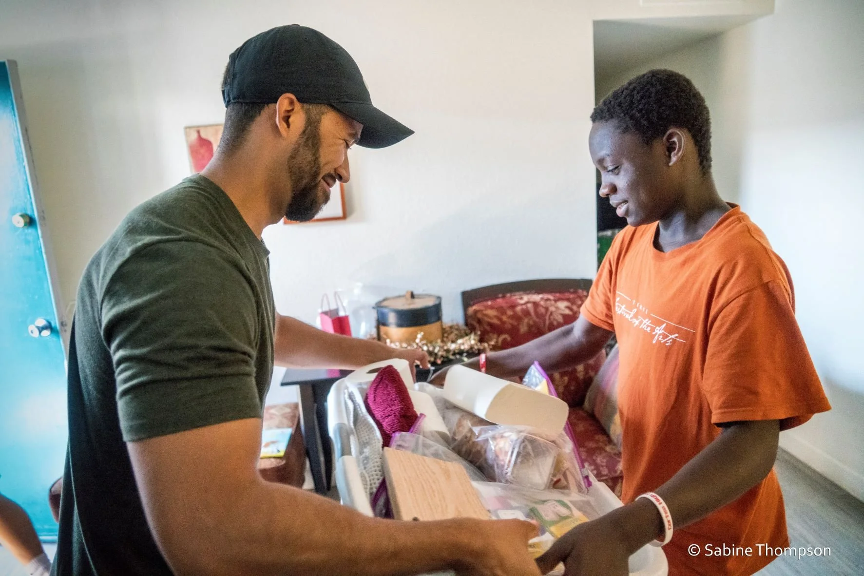 Two men exchanging gifts inside a living room, with one man receiving a box and the other handing it over, smiling.