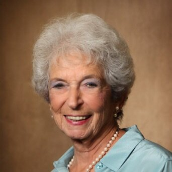 A smiling elderly woman with short, curly gray hair, wearing a light blue blouse and pearl necklace, against a plain brown background.