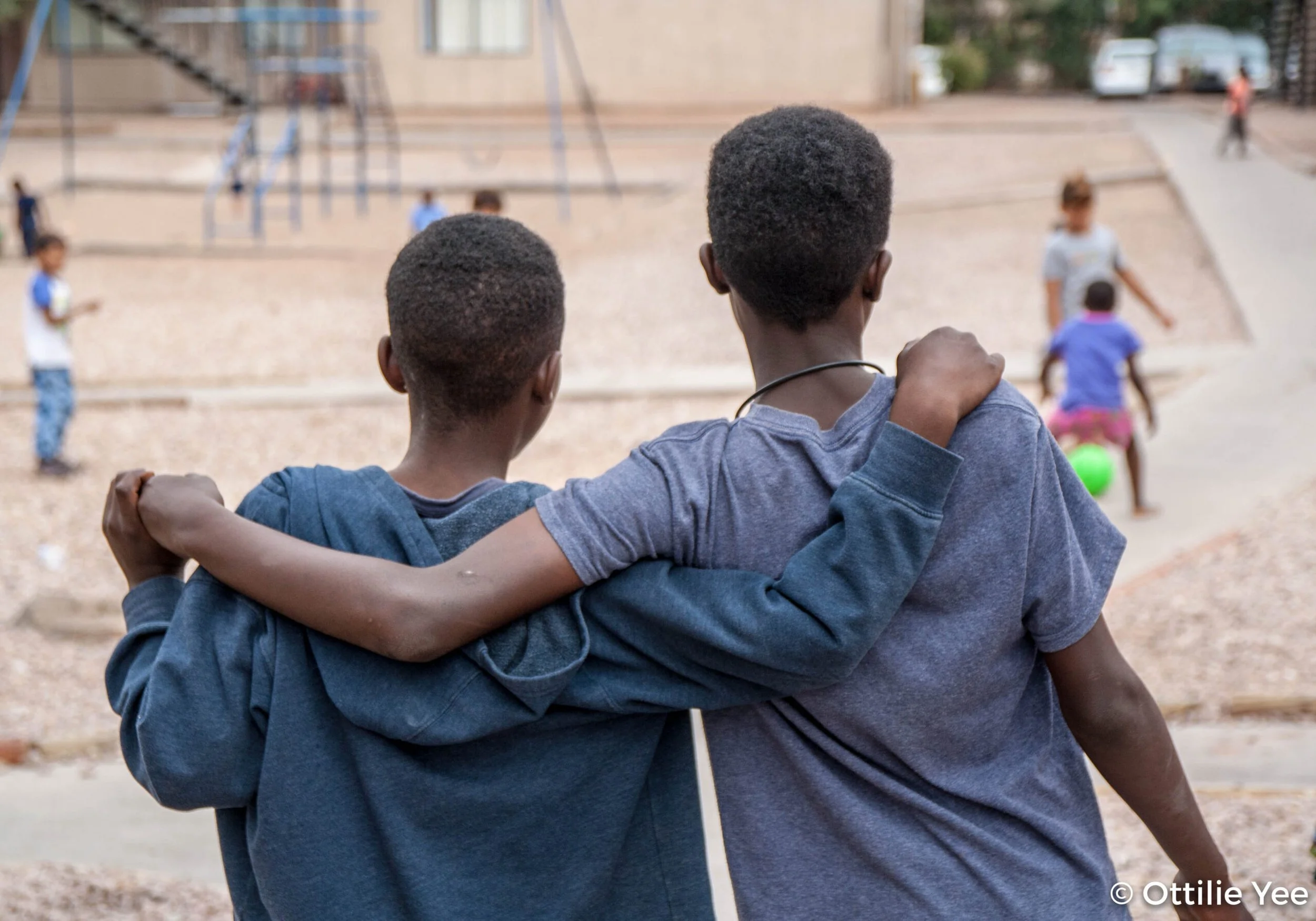 Two boys with their arms around each other's shoulders sitting on a ledge, overlooking children playing in a playground area.
