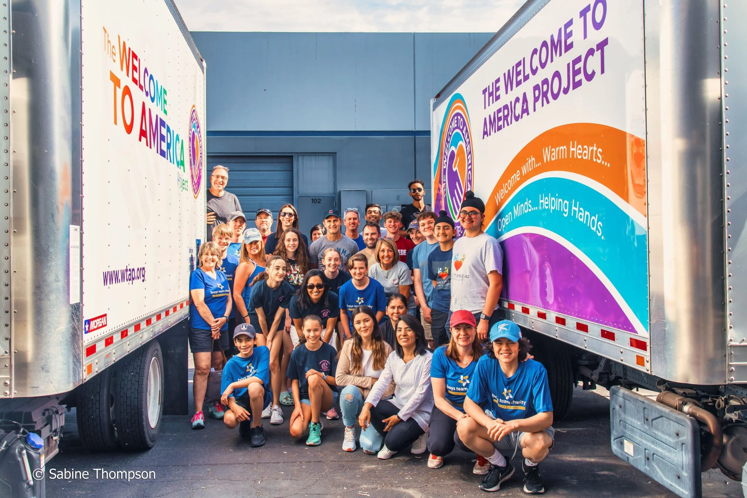 Group of people standing between two trucks with colorful 'Welcome to America' signs, smiling for a group photo outside warehouse.