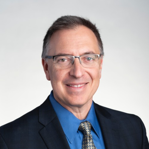 Professional headshot of a middle-aged man with glasses, short brown hair, wearing a blue shirt, patterned tie, and dark suit jacket, smiling at the camera against a light gray background.