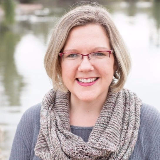 A smiling woman with shoulder-length blonde hair, red glasses, wearing a gray top and a knit scarf, standing outdoors near a body of water.