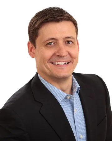 A smiling man in a dark suit jacket and light blue dress shirt against a white background.
