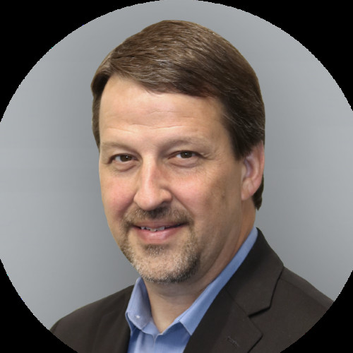 Portrait of a middle-aged man in a dark suit and blue shirt, smiling against a gray background.