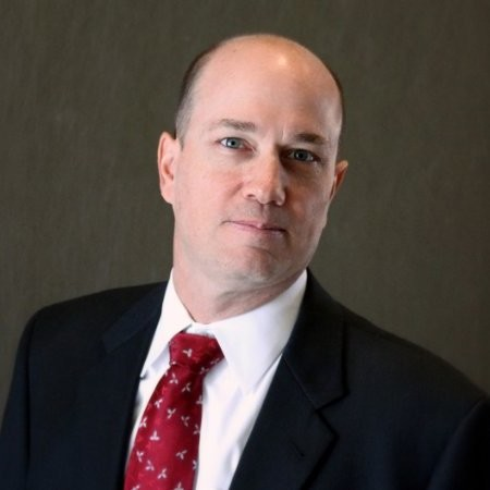 A professional headshot of a middle-aged man with short hair, wearing a dark suit, white shirt, and red tie, with a neutral background.