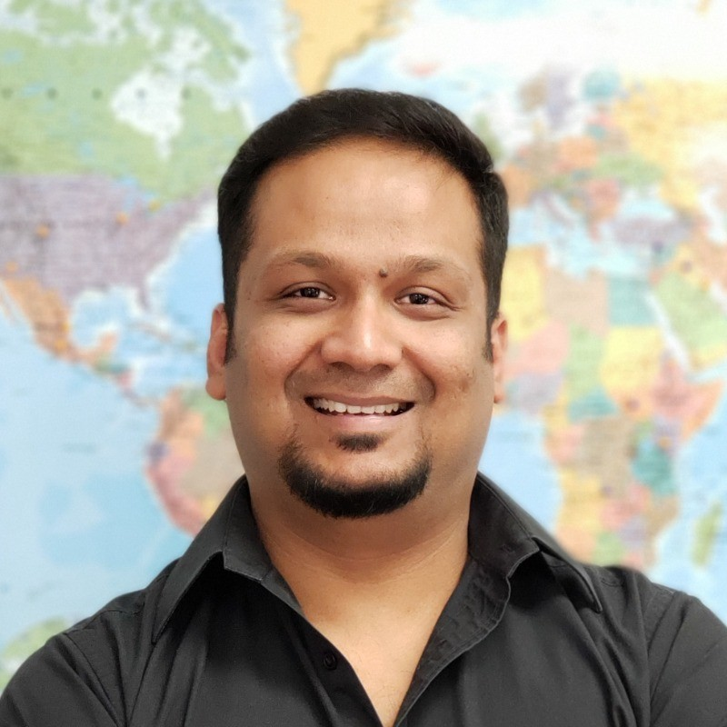 A man with short dark hair and a goatee smiling, standing in front of a world map.