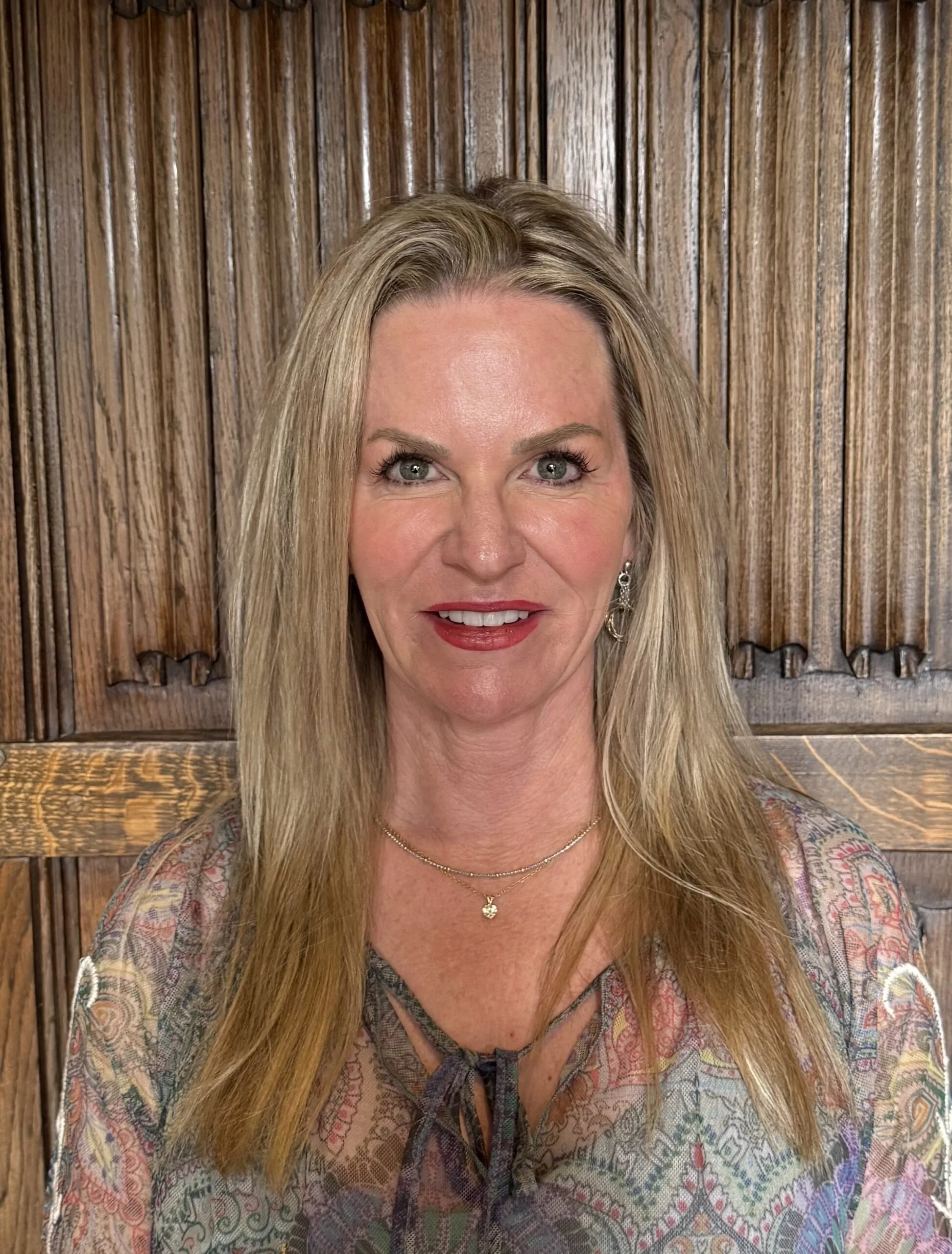 A woman with long blonde hair, wearing earrings, a necklace, and a patterned top, standing in front of a wooden wall.
