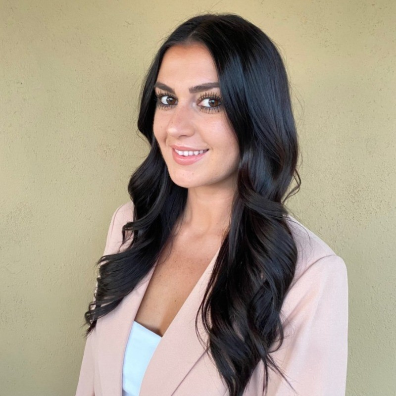 A woman with long, wavy black hair, smiling and wearing a light-colored blazer over a white top, standing against a plain beige wall.