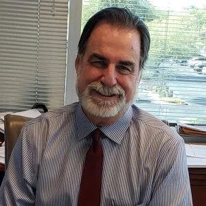 A smiling middle-aged man with dark hair and a beard, wearing a striped shirt and a red tie, sitting at a desk with blinds and a window in the background.