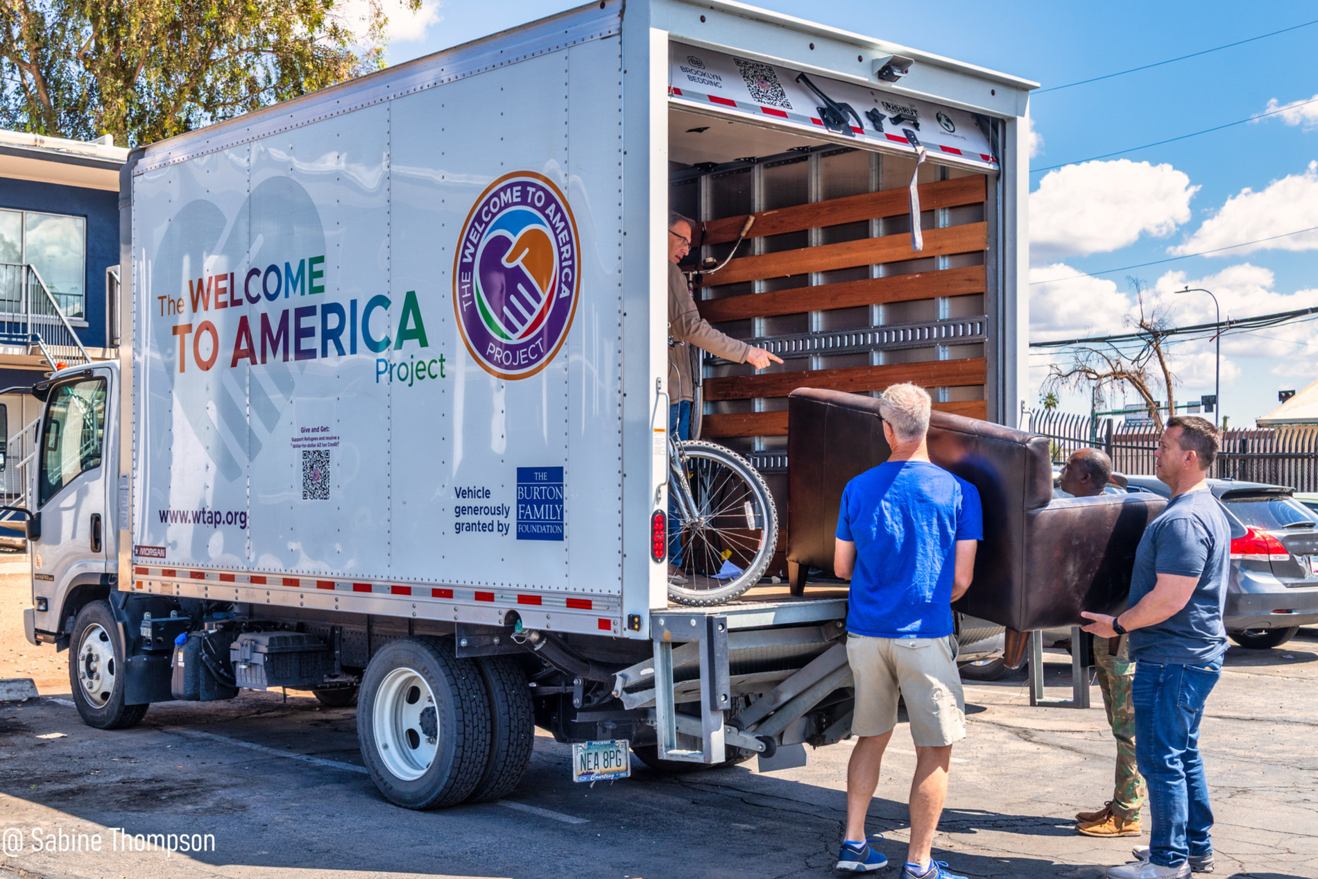 People unloading furniture from a truck with 'Welcome to America' project branding, parked in a lot with other cars and trees in the background.