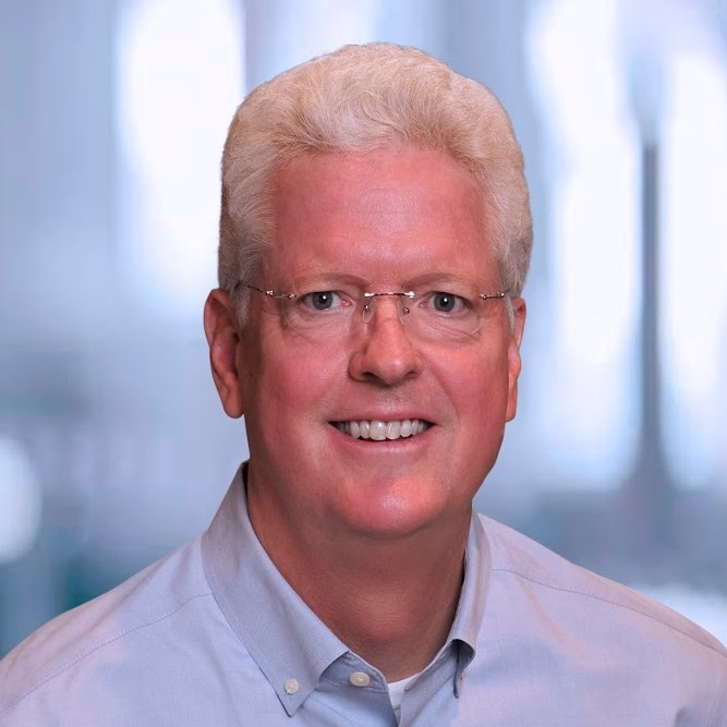 A middle-aged man with blonde hair, glasses, and a light blue shirt, smiling in an office setting.