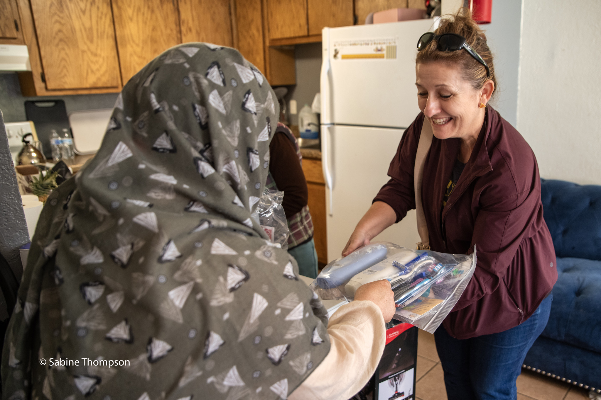Woman smiling while giving a plastic bag containing various items to a person wearing a hijab in a kitchen.