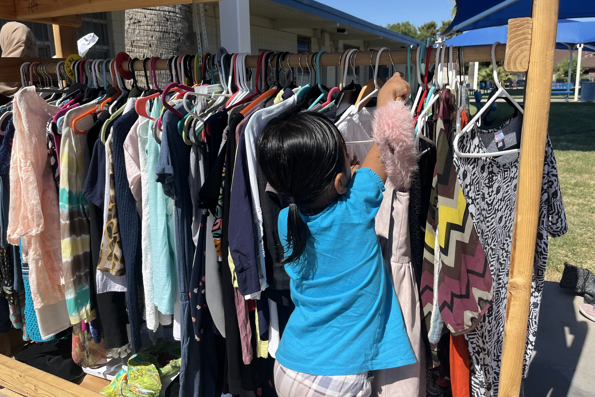A young girl with black hair in a ponytail, wearing a blue shirt and plaid shorts, shopping for clothes on a rack at an outdoor yard sale or market.