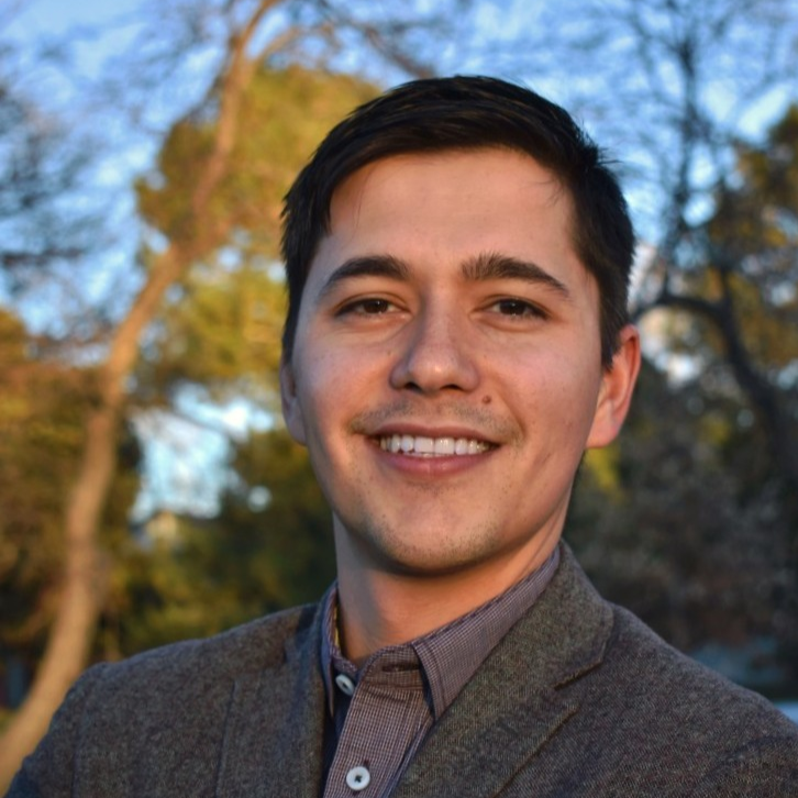 A smiling young man outdoors with trees and blue sky in the background, wearing a brown blazer and a button-up shirt.
