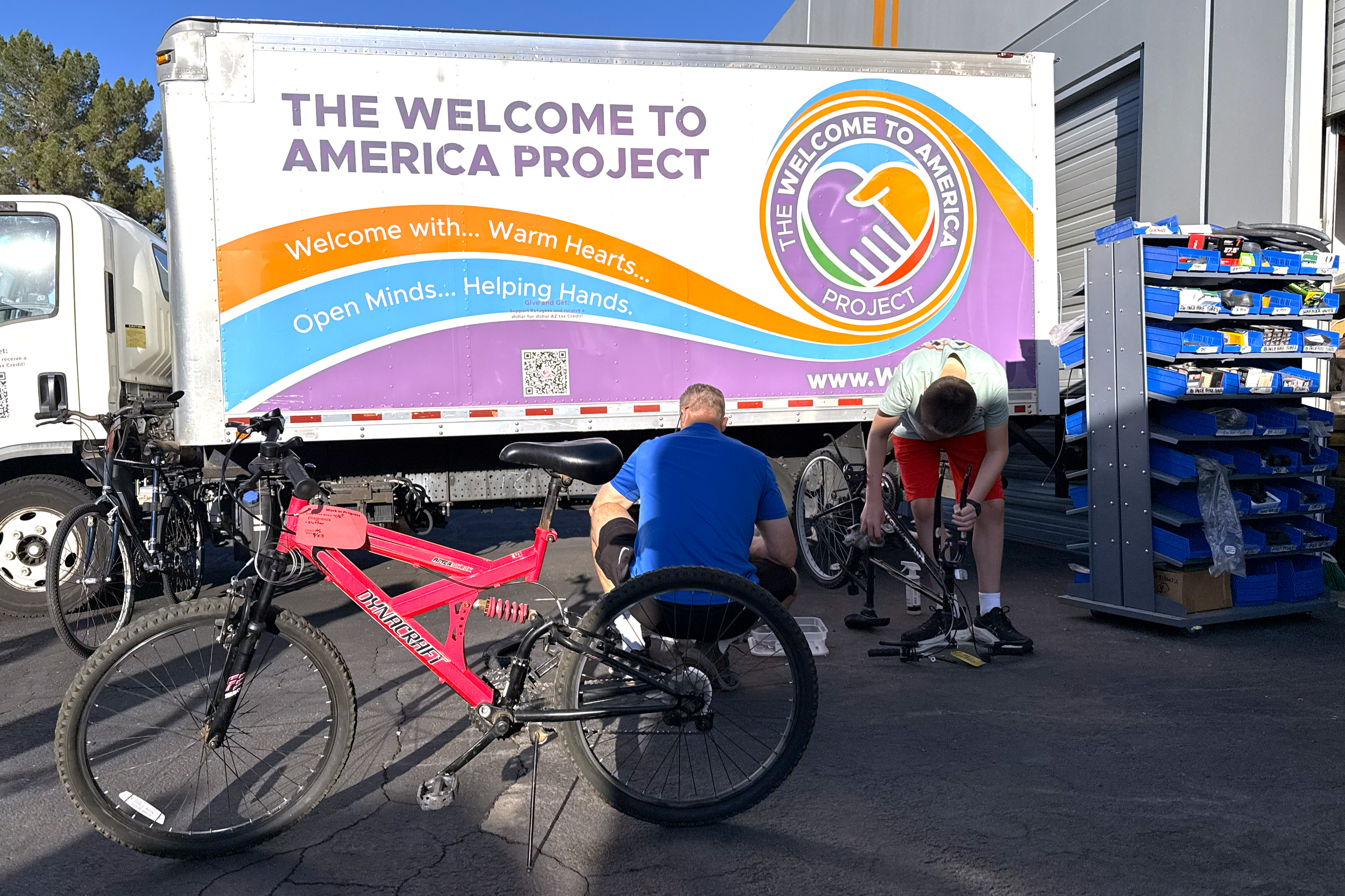 Two people working on bicycle repairs near a truck with a colorful banner that reads "The Welcome to America Project" and "Welcome with.. Warm Hearts... Open Minds... Helping Hands." A red bicycle is in the foreground, and blue storage bins are on a shelf nearby.