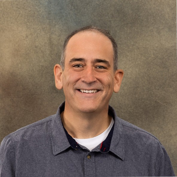 A smiling man wearing a gray button-up shirt in front of a textured beige background.