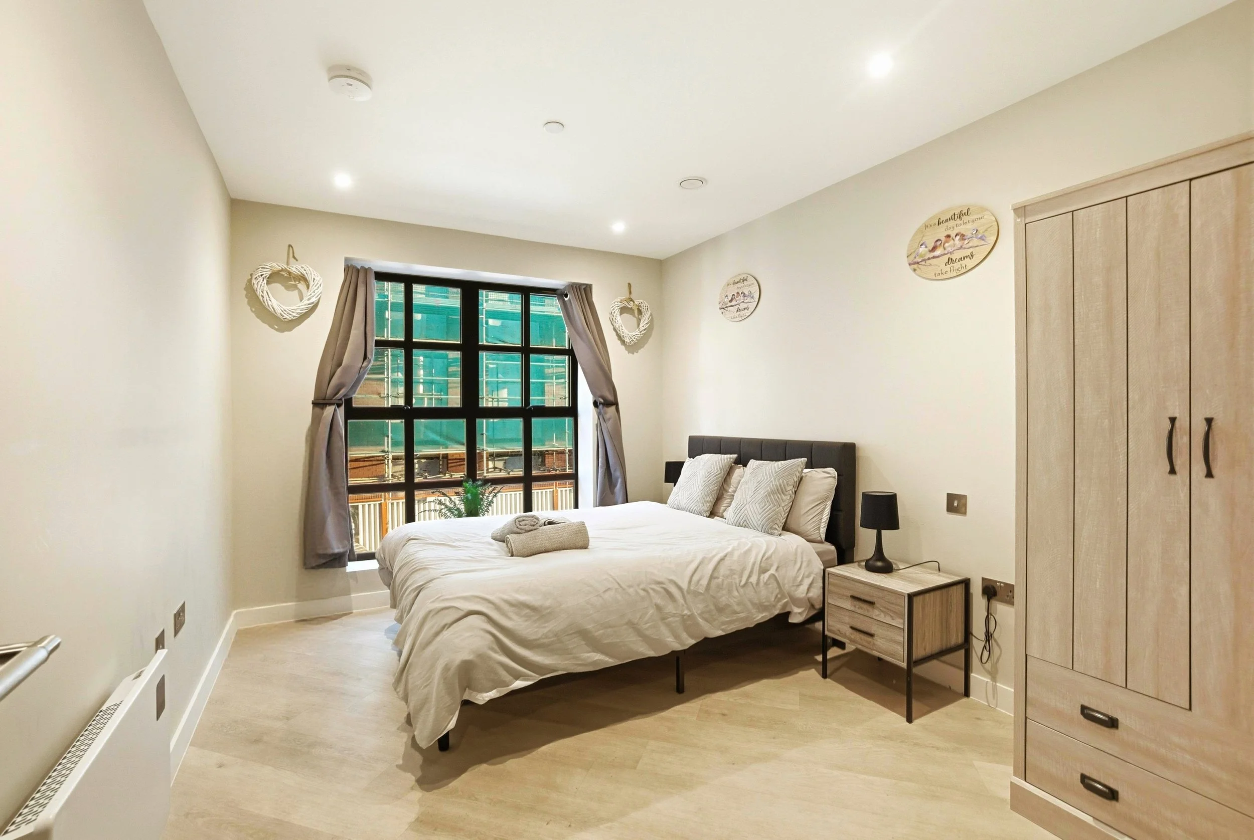 Bedroom with a bed, pillows, bedside table with a lamp, a wardrobe, window with curtains, wall decorations, and a towel on the bed.