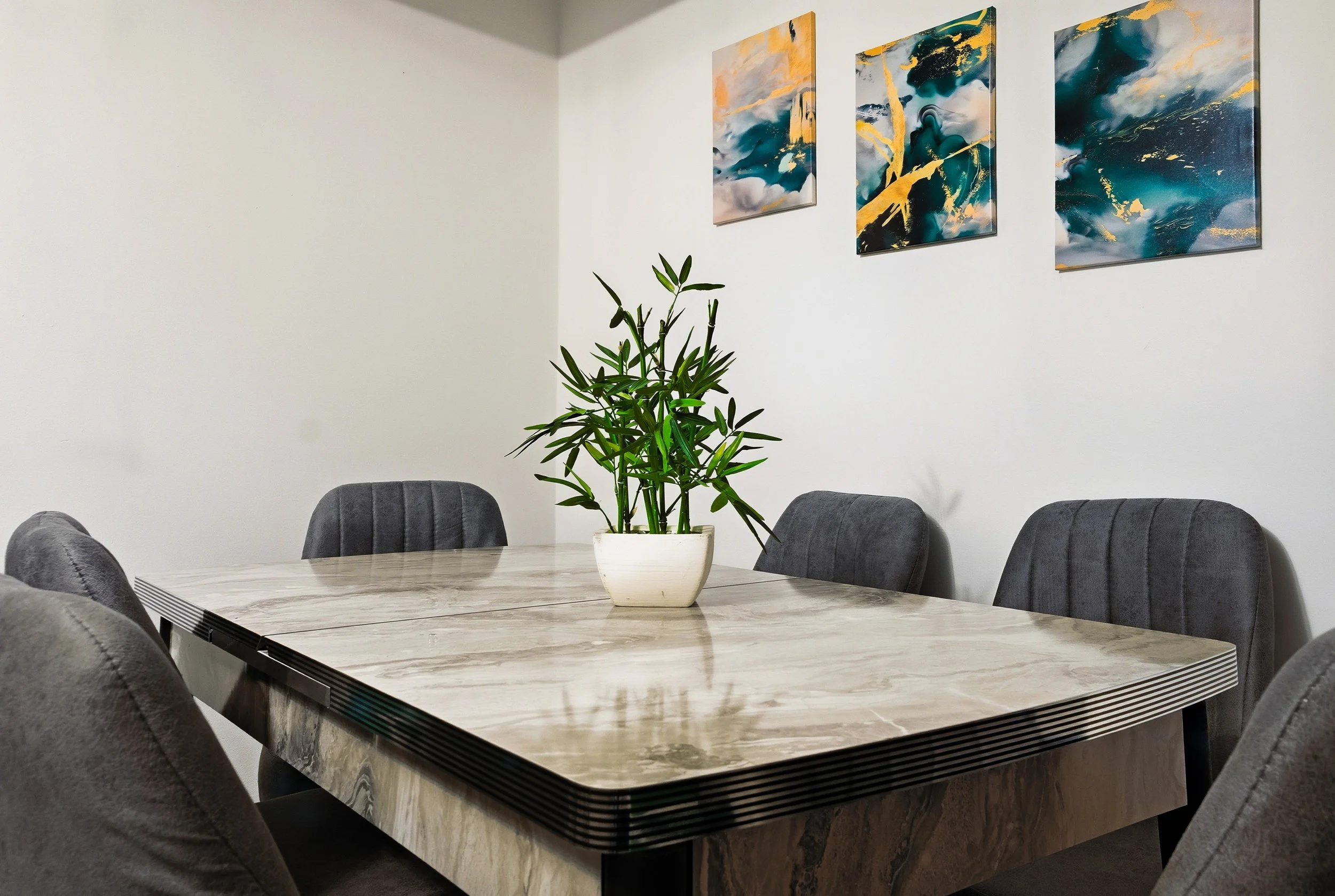 A modern conference room with a marble table, six gray upholstered chairs, a potted plant in the center, and abstract artwork on the white wall.