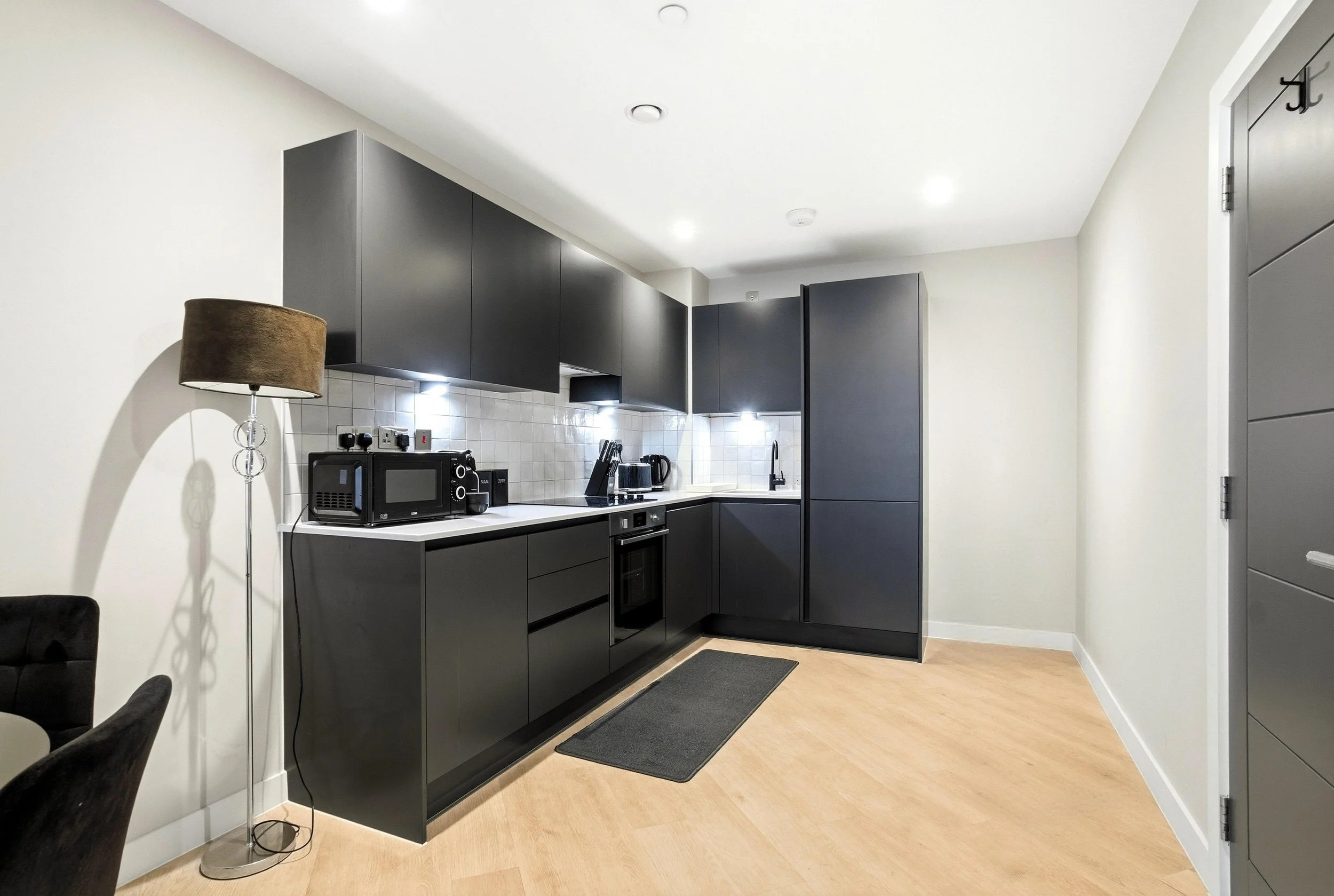 Modern kitchen with black cabinets, white backsplash, and wooden flooring, including appliances like microwave, toaster, and kettle. There is a tall black pantry and a small gray rug on the floor.
