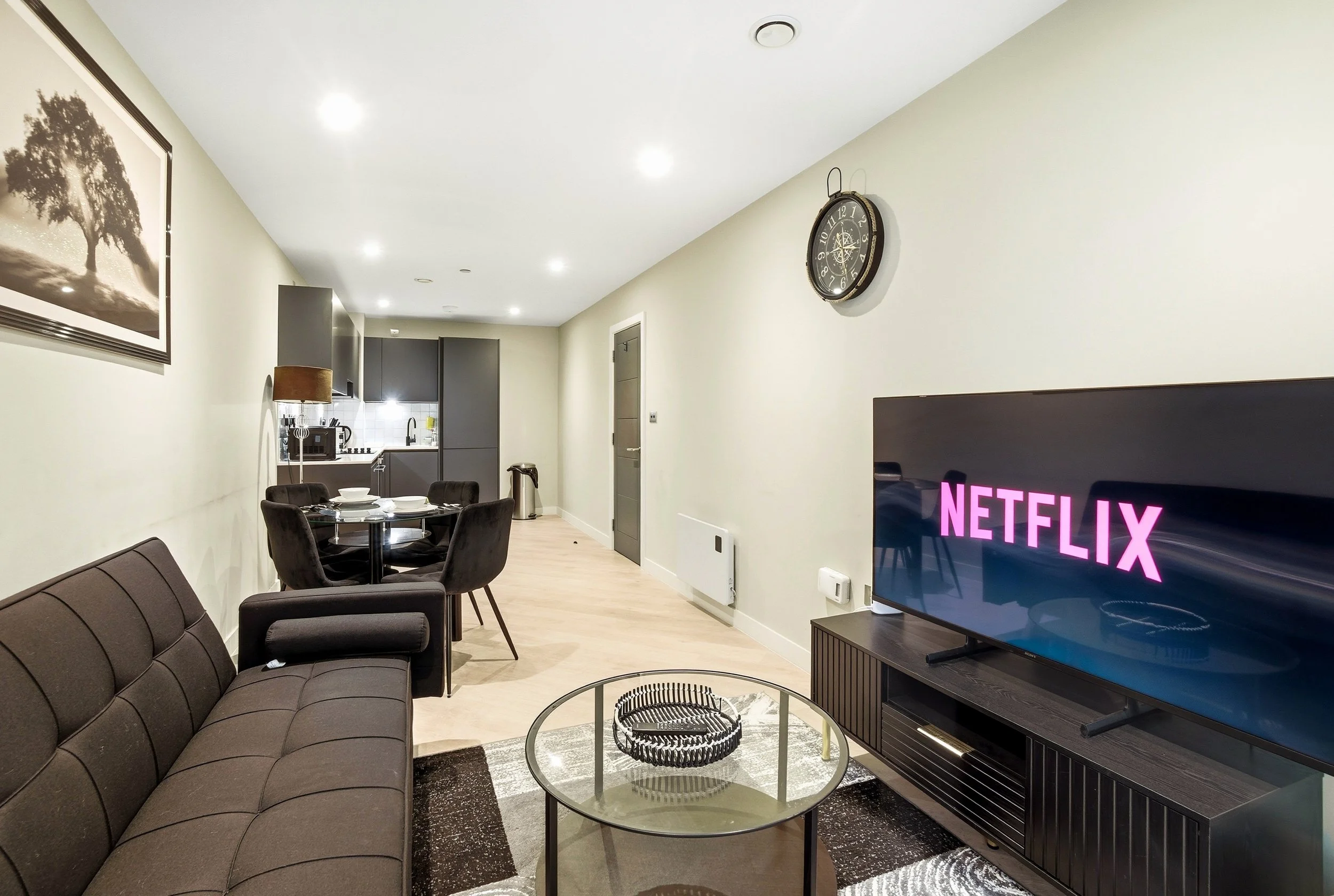 Modern living room with brown sofa, black round coffee table, and TV displaying Netflix logo, with dining area and kitchenette in the background.