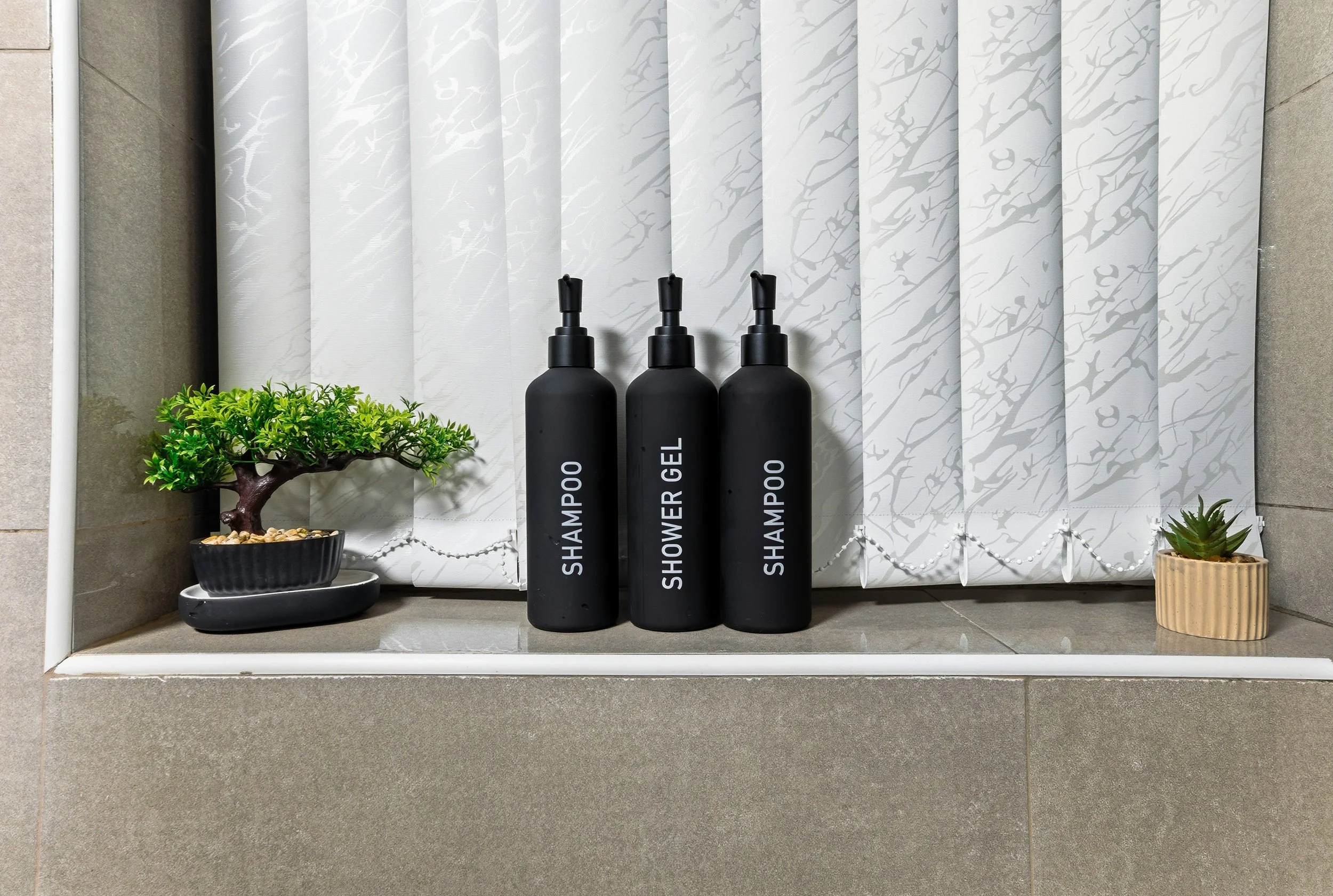 Shampoo, shower gel, and shampoo bottles on a bathroom shelf with a small bonsai tree and a potted succulent plant.