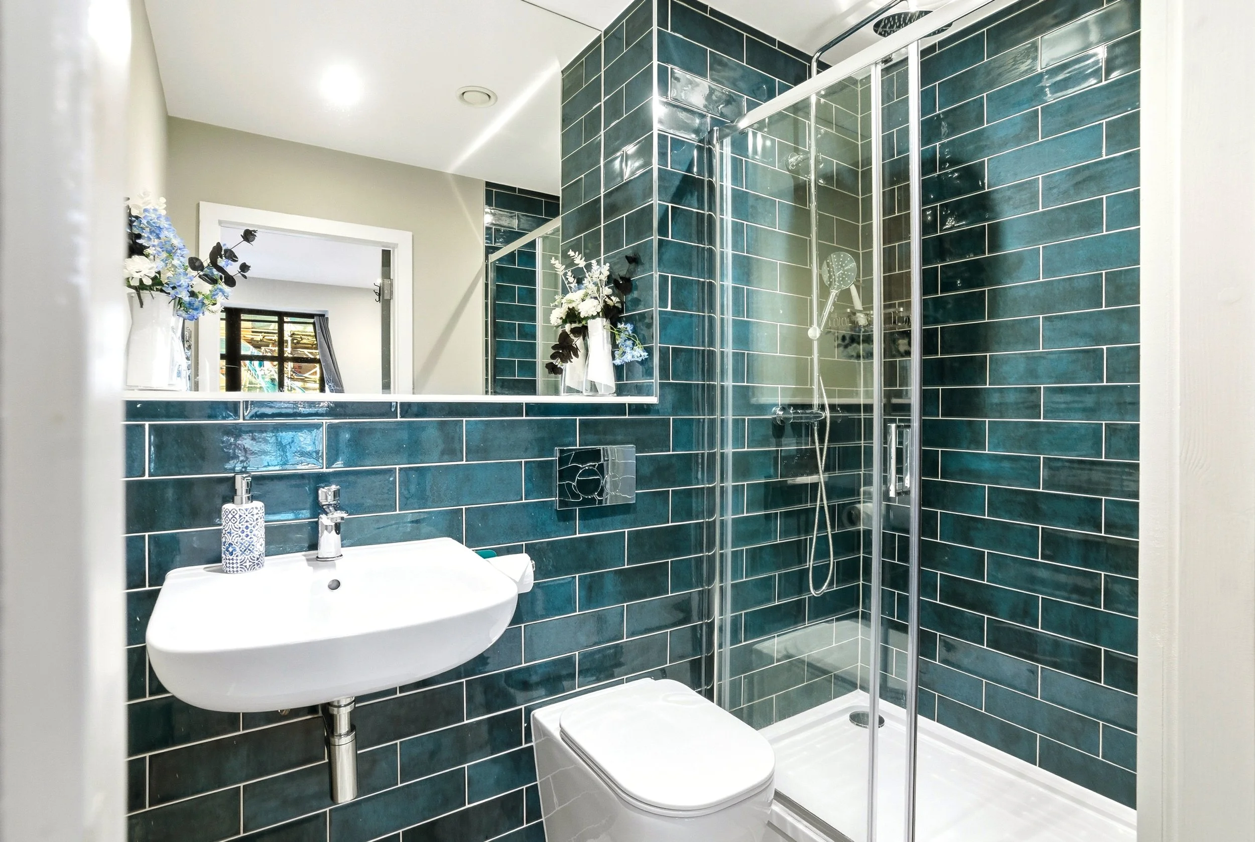 A modern bathroom with teal subway tile on the walls, a white sink with a blue patterned soap dispenser, a mirror, a small window with a black curtain, and a glass shower enclosure with a rainfall showerhead and a hand shower.