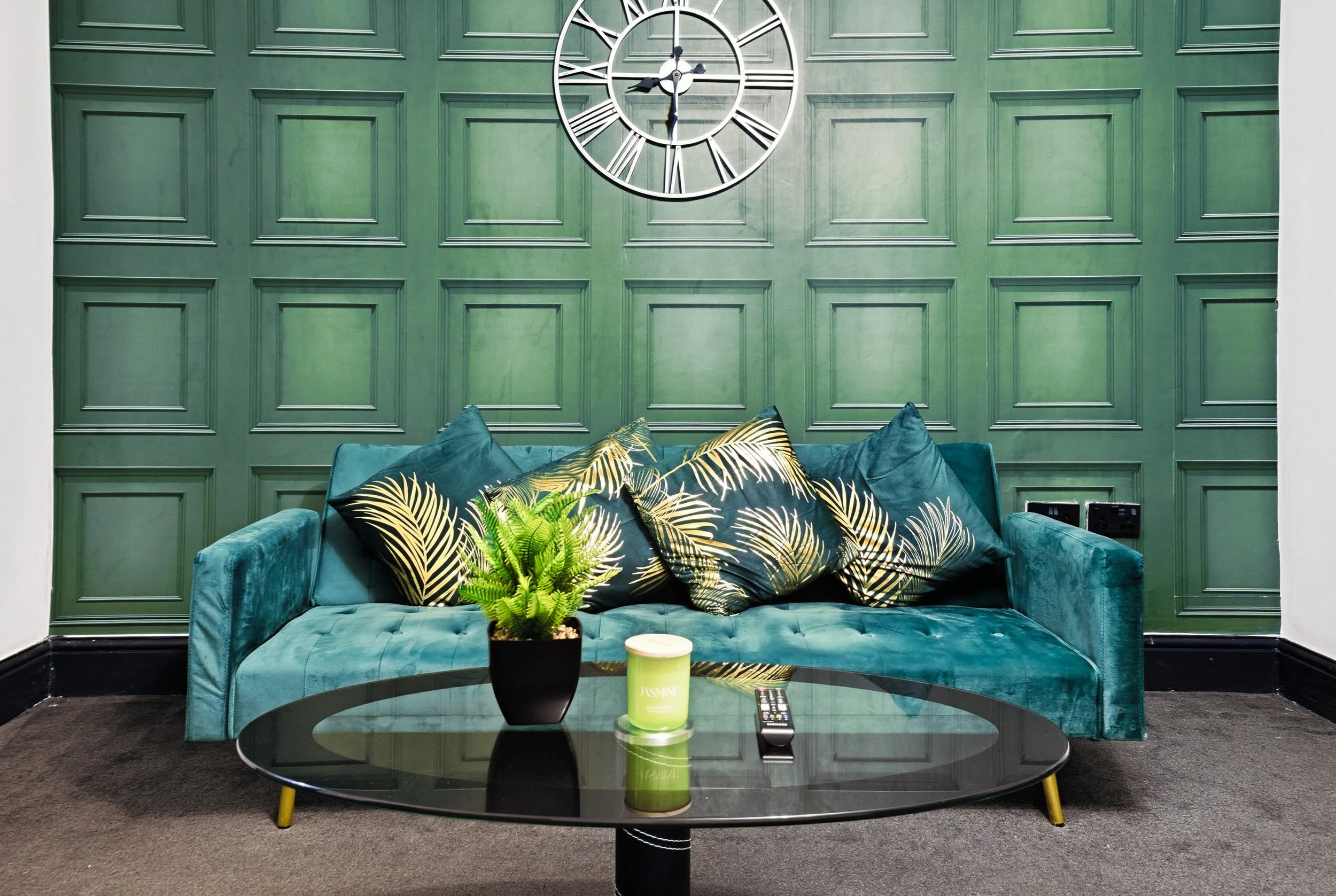A teal velvet couch with five decorative pillows, three with black and gold leafy patterns, on a gray carpeted floor against a green paneled wall. A black coffee table with a potted fern, a green candle, and a TV remote is in front of the couch. A large circular wall clock with Roman numerals is mounted on the wall above the couch.