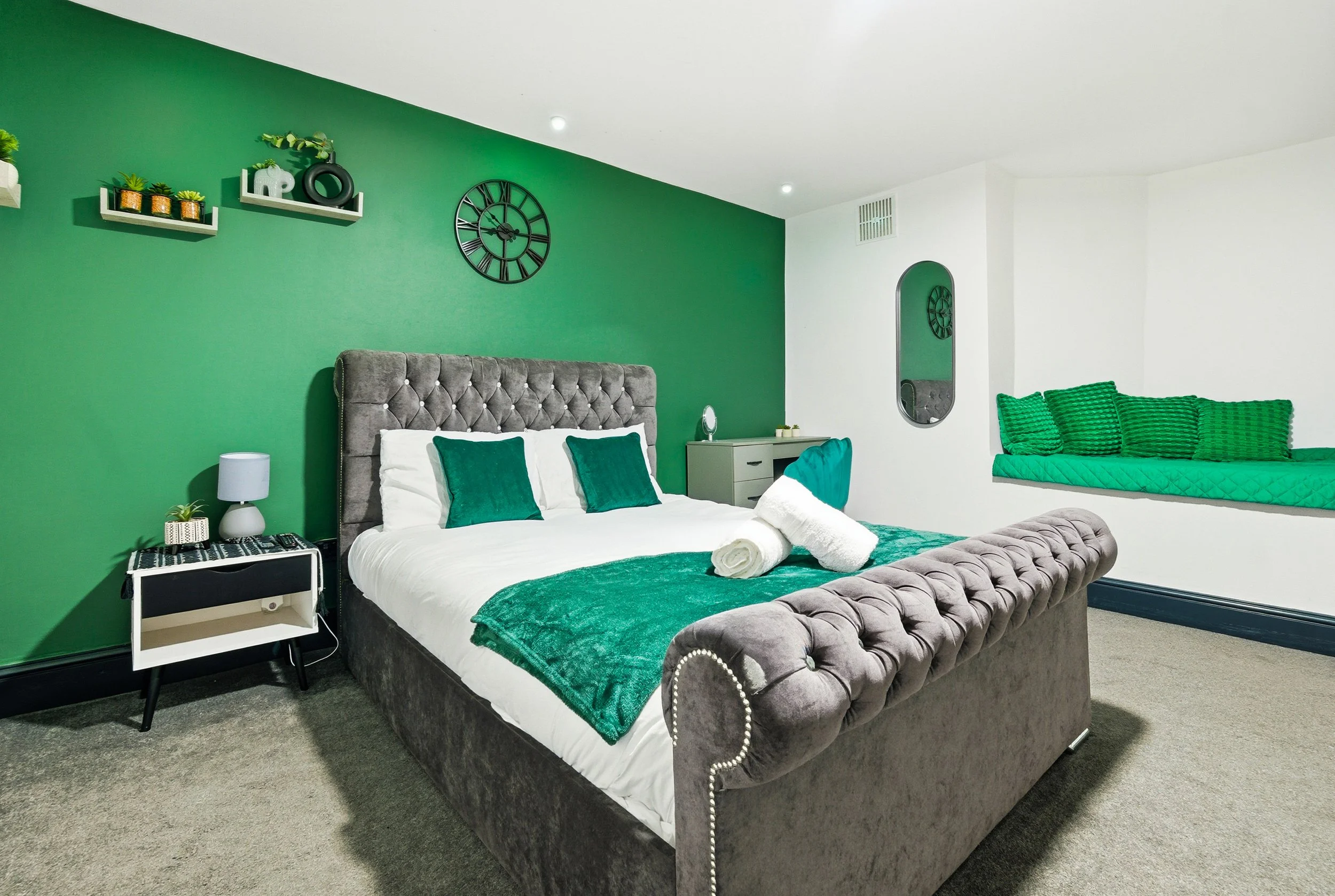 A bedroom with a green accent wall behind a gray tufted bed, white bedding, green blankets, and matching pillows. A small white nightstand with a lamp and plant is beside the bed. A gray desk with a mirror and other items is across from the bed. Ther