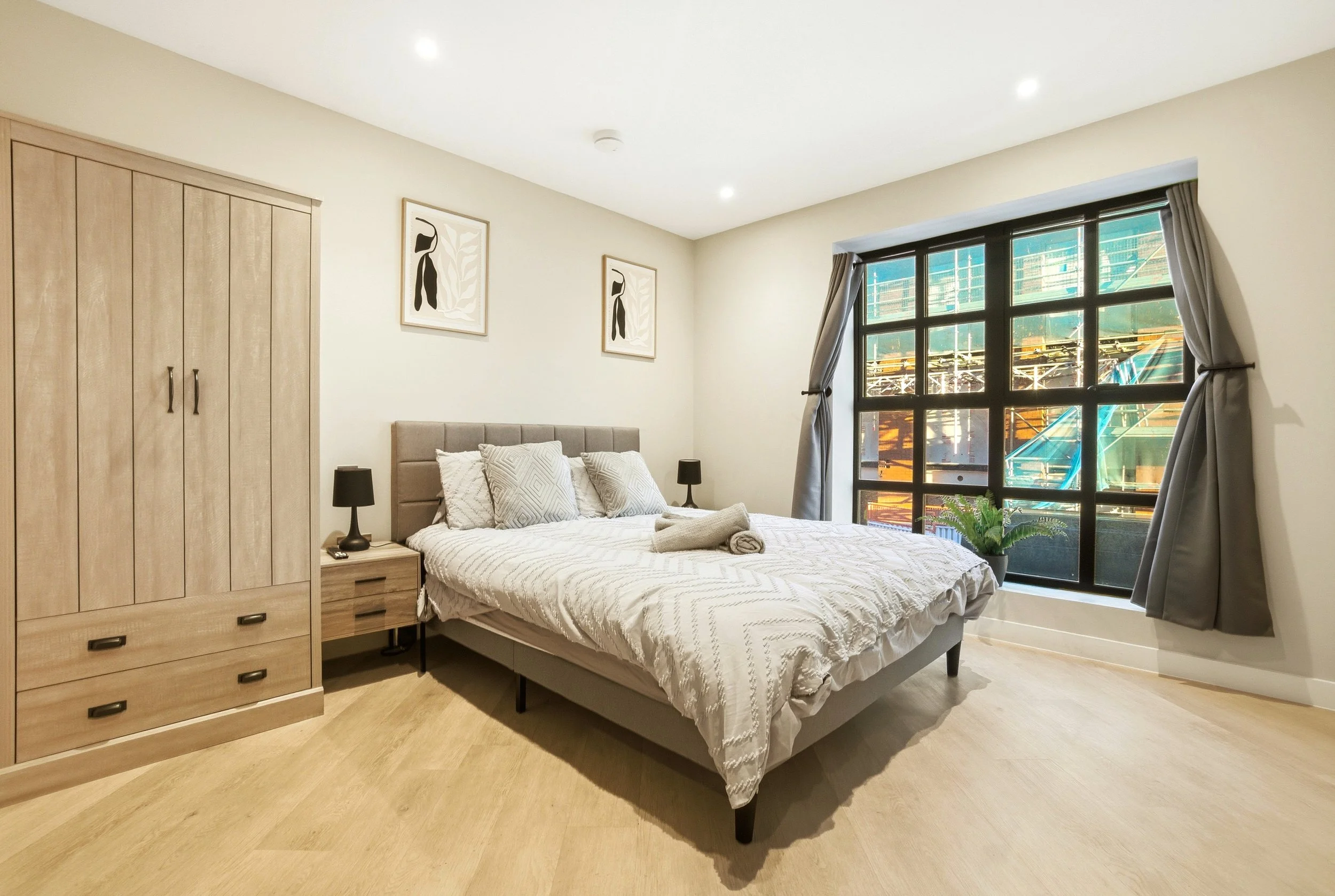A modern bedroom with a large bed, wooden wardrobe, and a big window with gray curtains, overlooking a construction site.