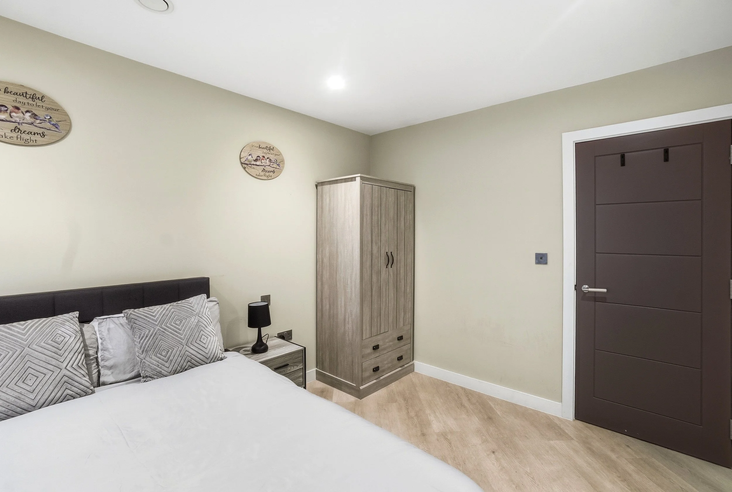 A bedroom with a bed, a small bedside table with a black lamp, a wardrobe, light-colored walls, a dark door, and decorative wall plaques.