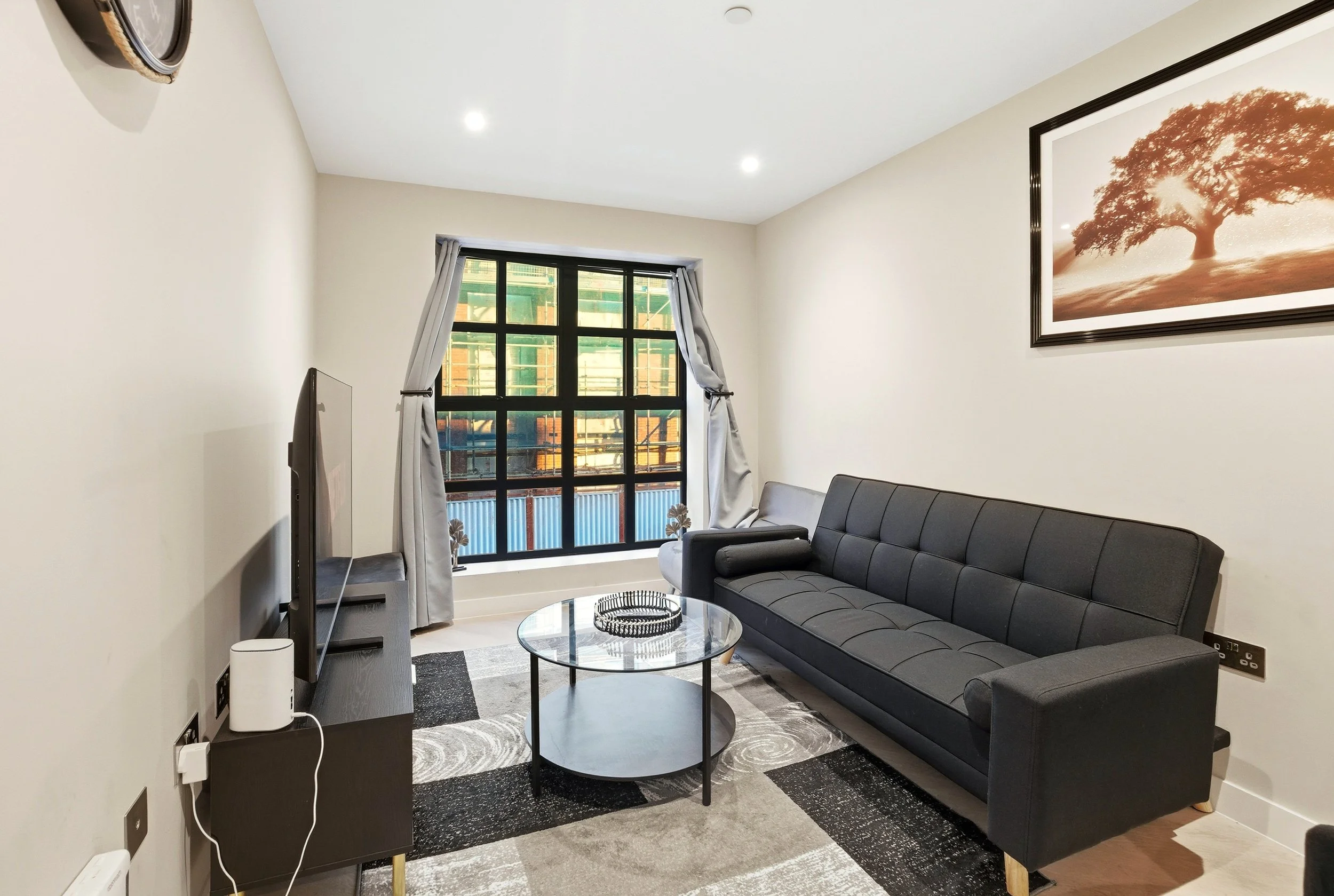 Living room with a large window, gray curtains, a black sofa, a glass coffee table, a framed picture of a tree on the wall, and a television on a black stand.