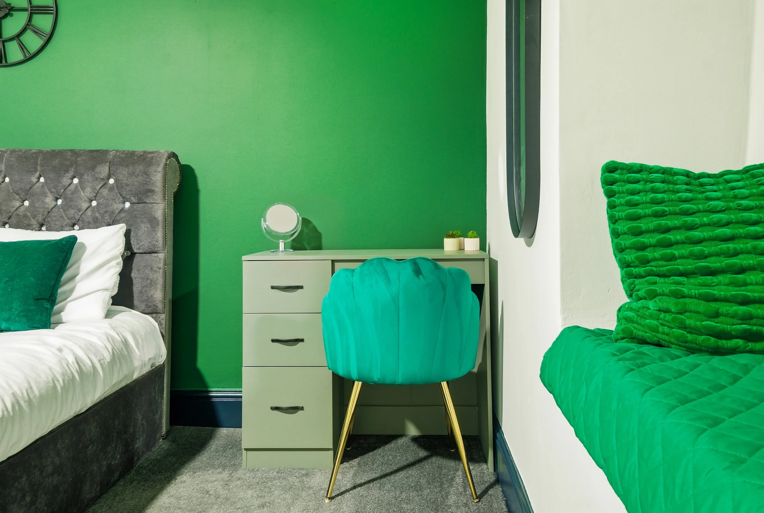Bedroom with green accents, grey bed with velvet headboard, green pillow, white bedsheets, green wall, desk with small mirror and plants, green shell-shaped chair, green textured sofa with cushions, and wall clock.