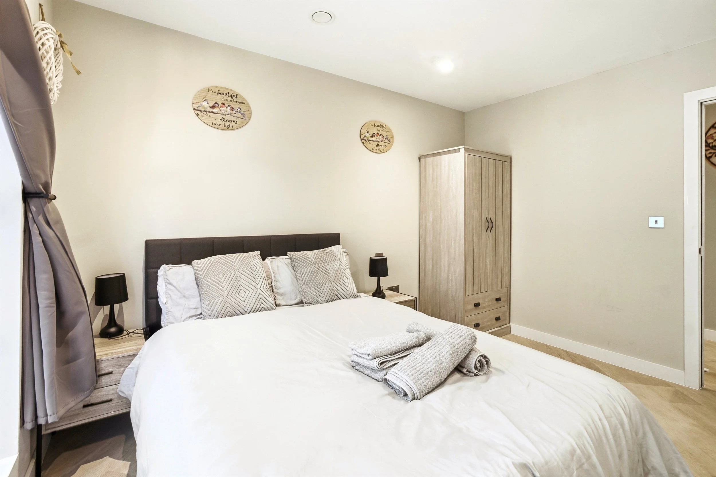 A neatly made bed with white sheets and four patterned pillows in a bedroom. Two black bedside lamps sit on matching nightstands. On the wall above the bed, there are two decorative plaques with motivational quotes. To the right, a wooden wardrobe wi