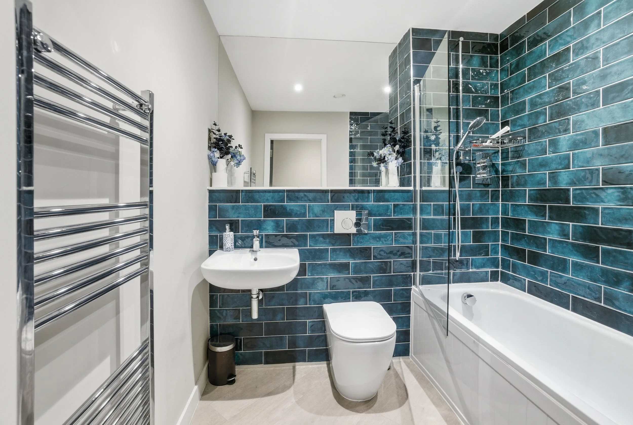 Modern bathroom with white fixtures and blue tiled walls, including bathtub, walk-in shower, toilet, small sink, and a wall-mounted towel radiator.