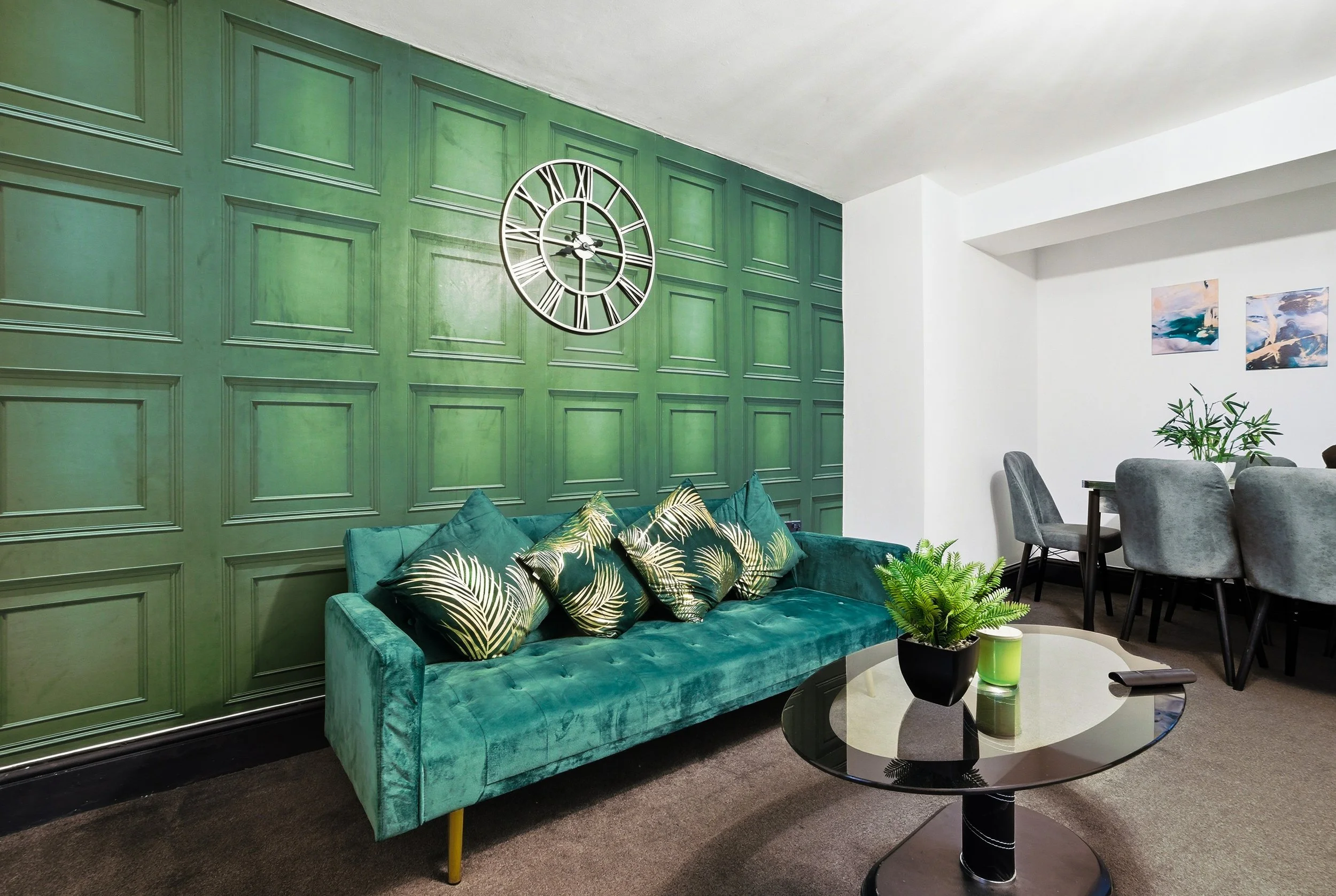 Living room with green paneled accent wall, teal sofa with patterned pillows, black glossy coffee table with potted plant, and dining area with gray chairs and artwork on white wall.