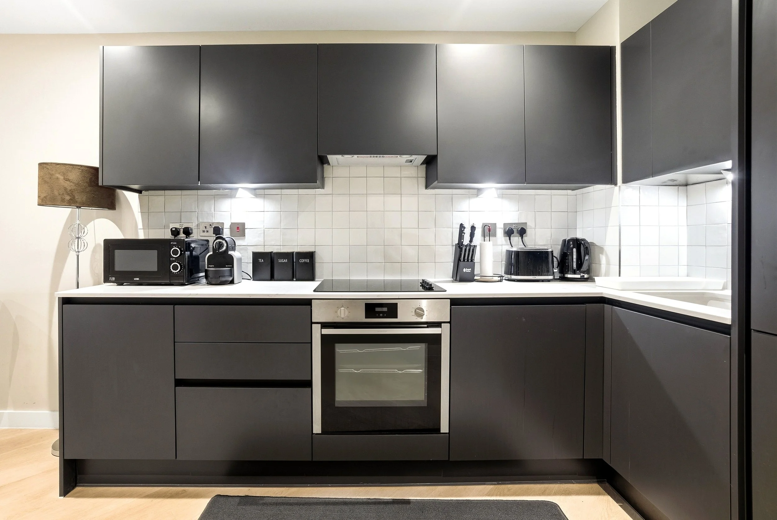 Modern kitchen with black cabinets, white countertop, oven, microwave, coffee maker, toaster, and knives, with a black rug on a wooden floor.