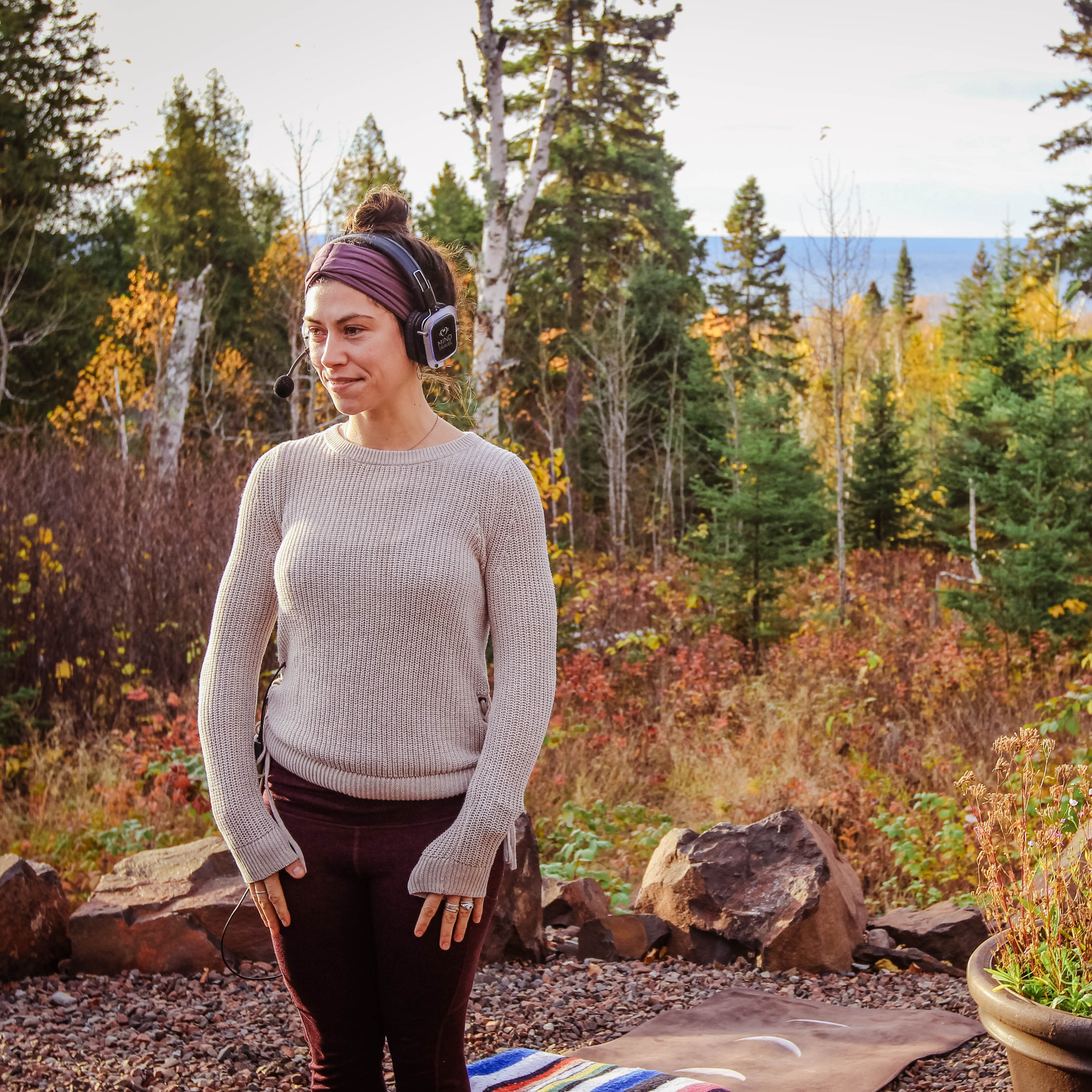 A woman standing outdoors among trees during autumn, wearing a beige sweater, black pants, and a purple headband, with a walkie-talkie headset on her head.