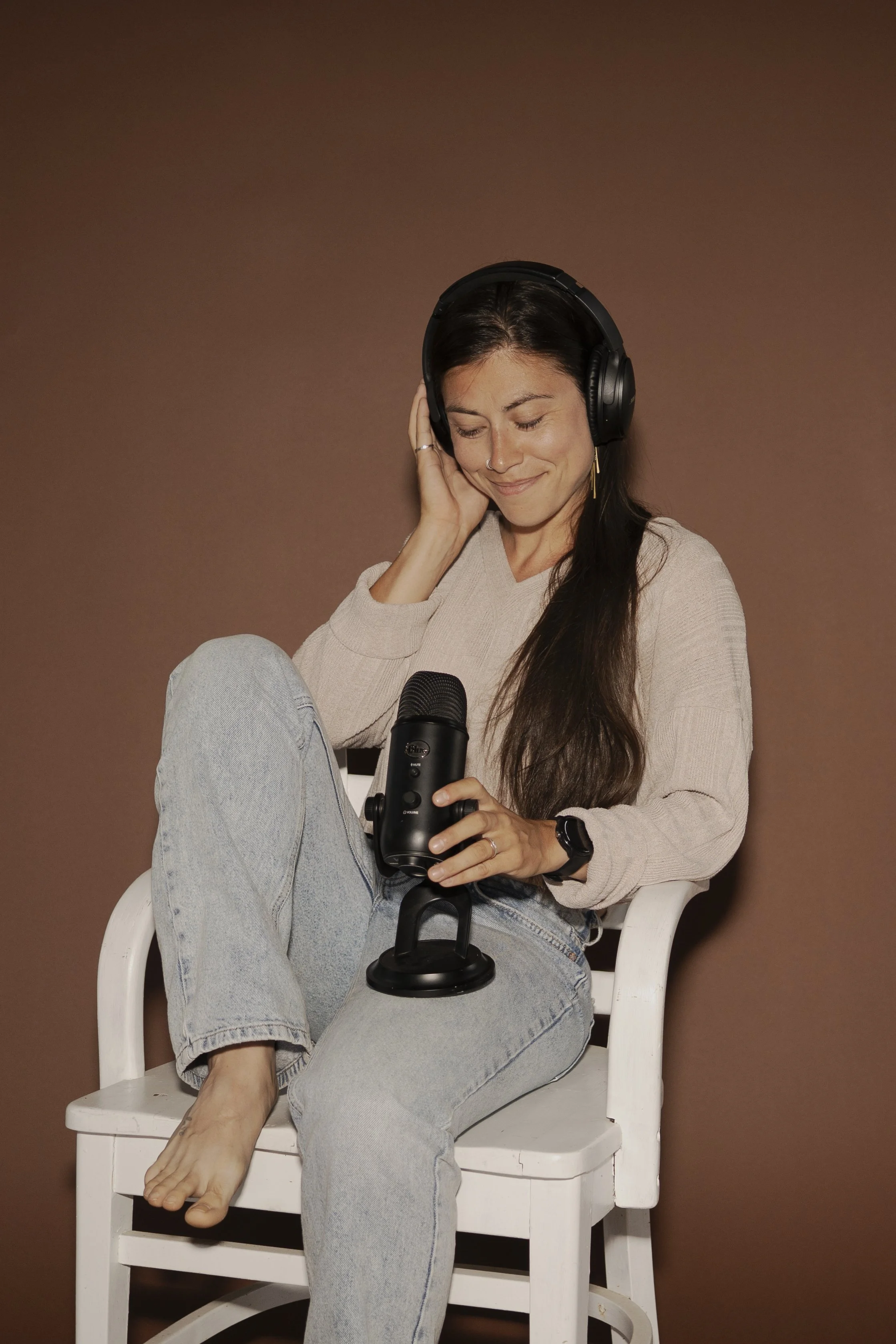 A woman with long dark hair, wearing a beige sweater, light wash jeans, and a wristwatch, sitting on a white chair against a brown background. She is smiling with eyes closed, wearing large black headphones, holding a microphone in her right hand, and resting her left hand on her cheek.