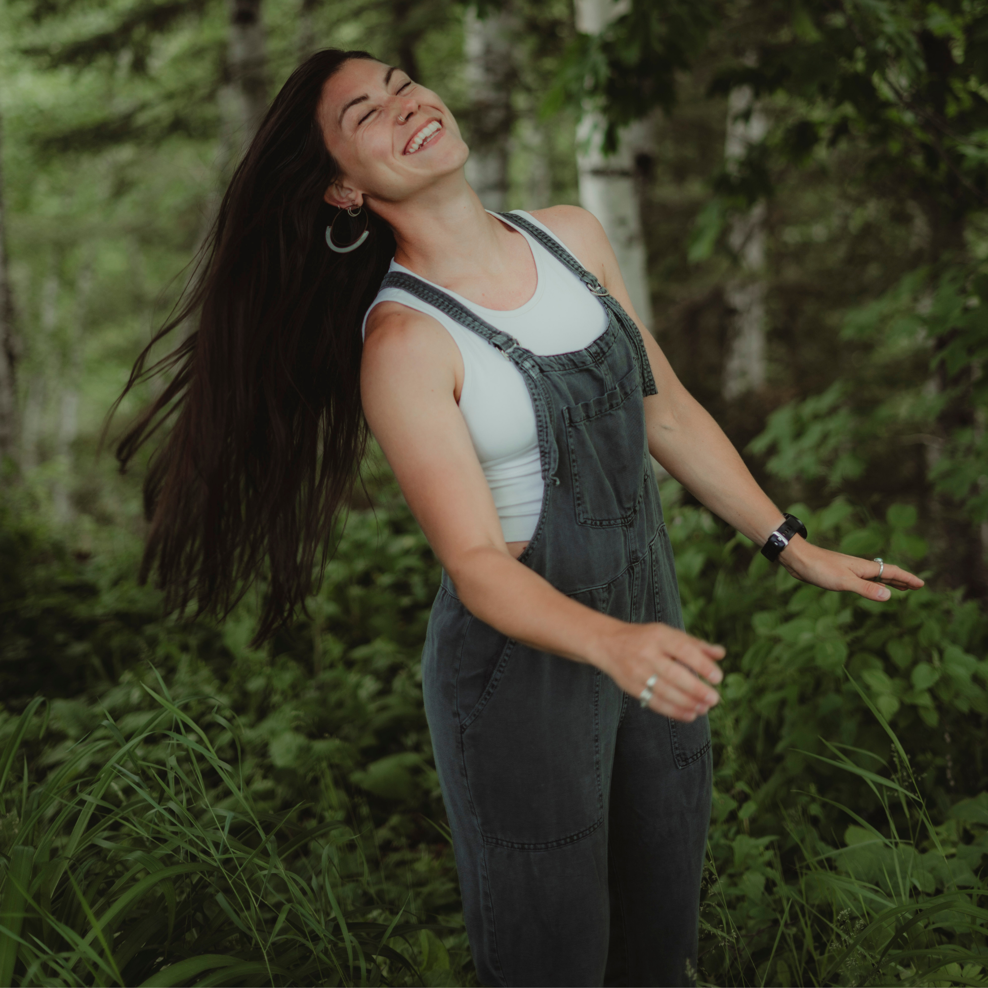 A woman with long dark hair smiling with her eyes closed outdoors among green trees and foliage.