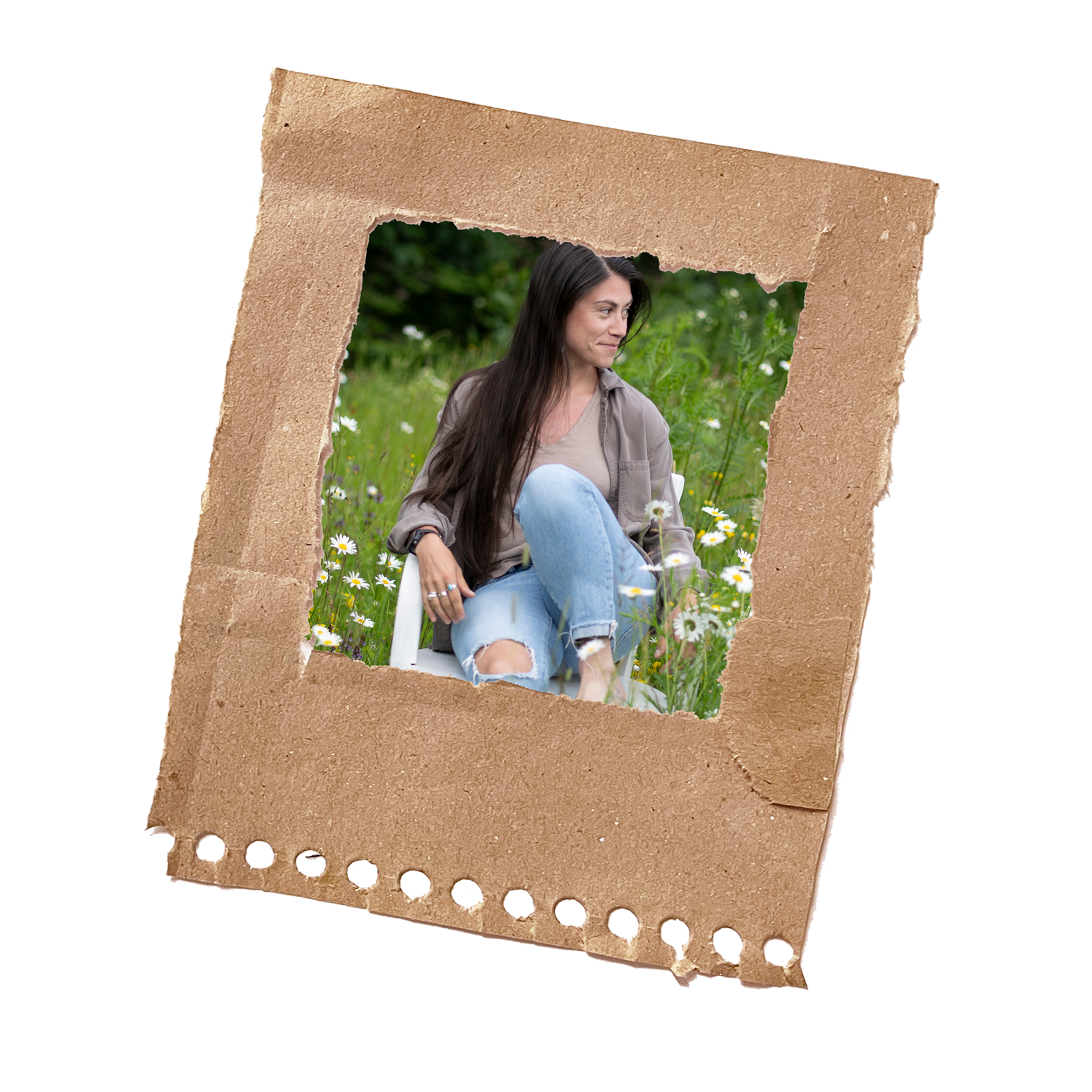 A woman sitting outdoors in a green field with white flowers, smiling and looking to the side, framed by a torn piece of brown cardboard.
