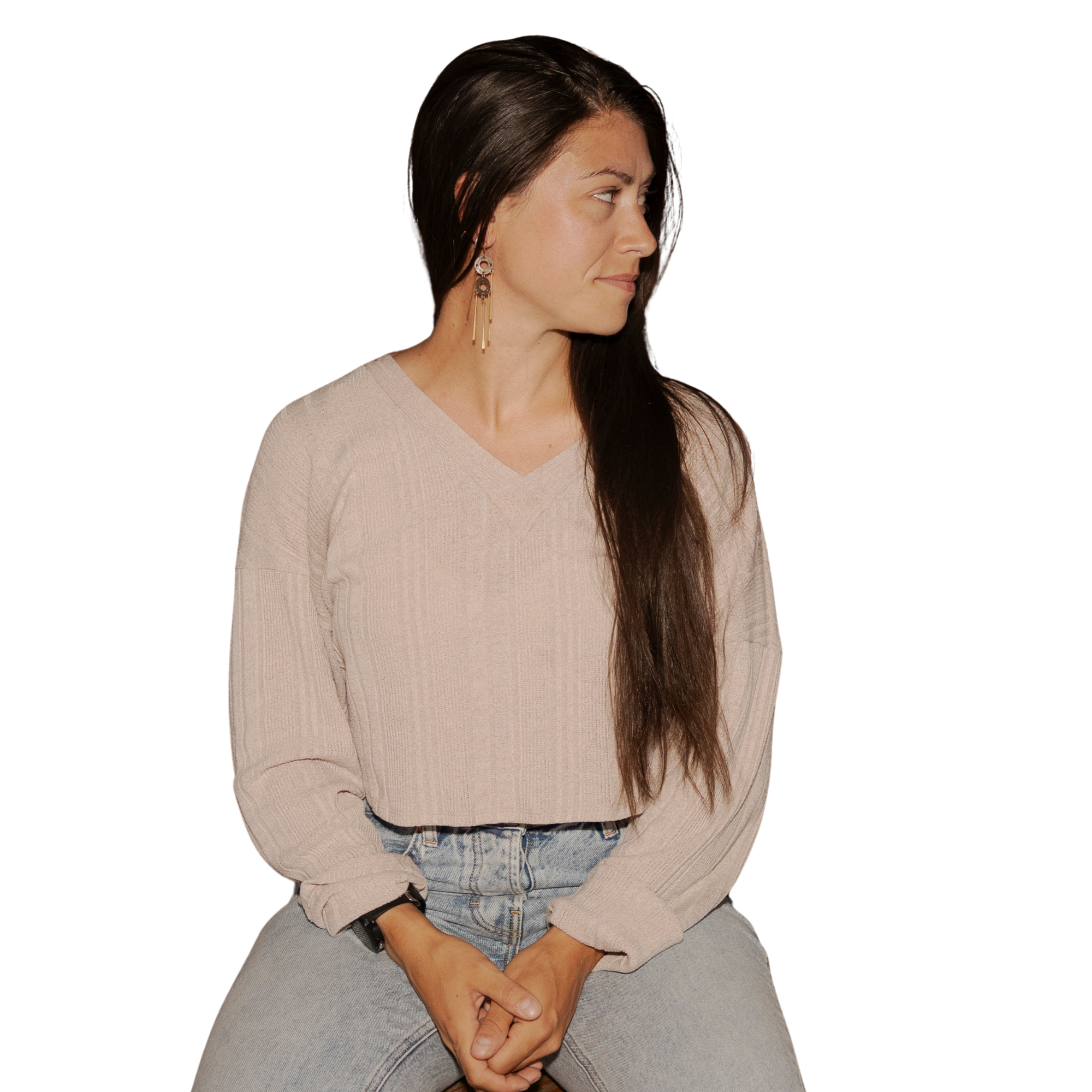 A woman with long dark hair wearing a beige sweater and blue jeans, sitting with her hands clasped, looking to her right, against a plain background.