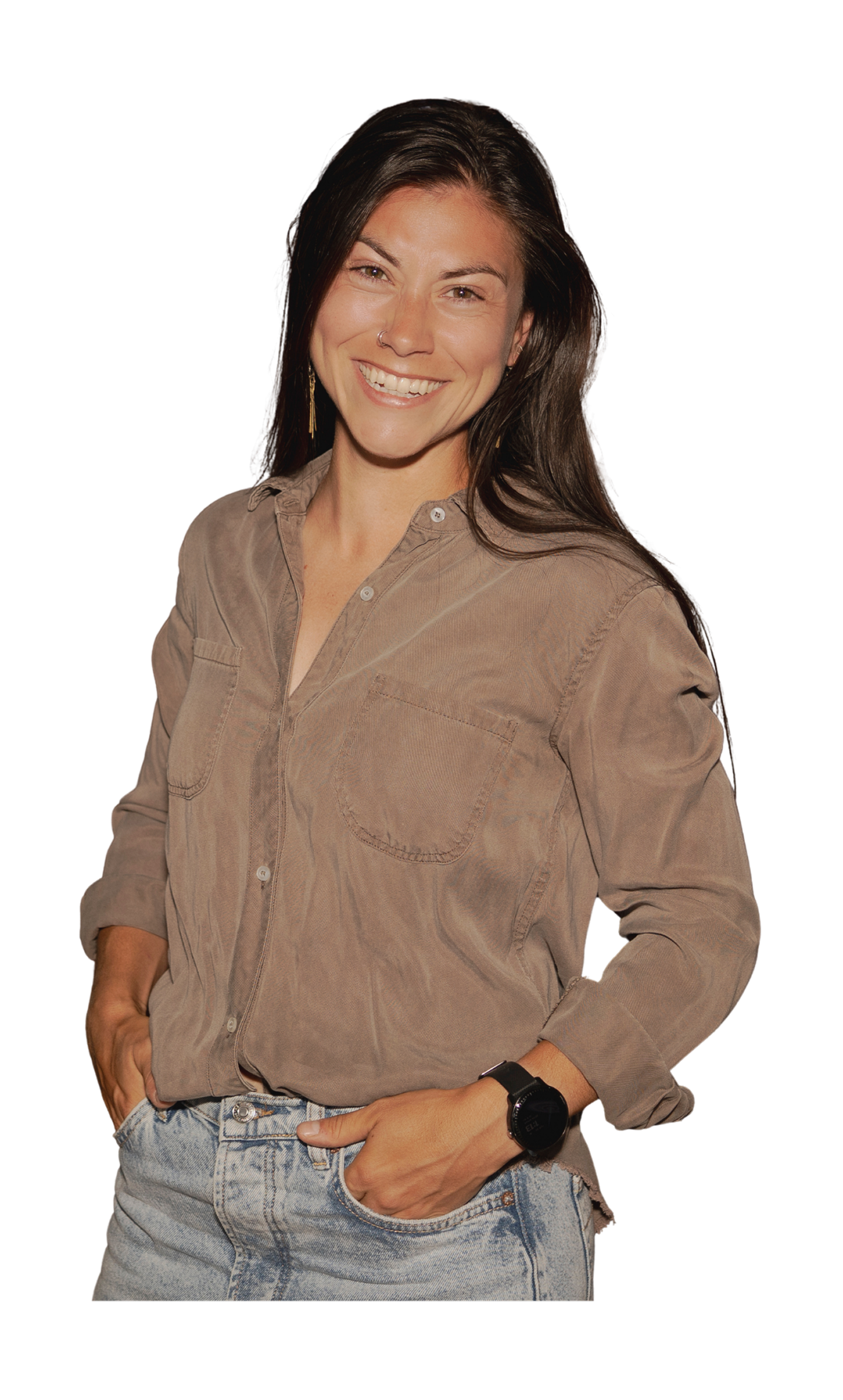 Young woman with long dark hair smiling, wearing a beige button-up shirt, jeans, and a black watch, standing with one hand in her pocket against a black background.