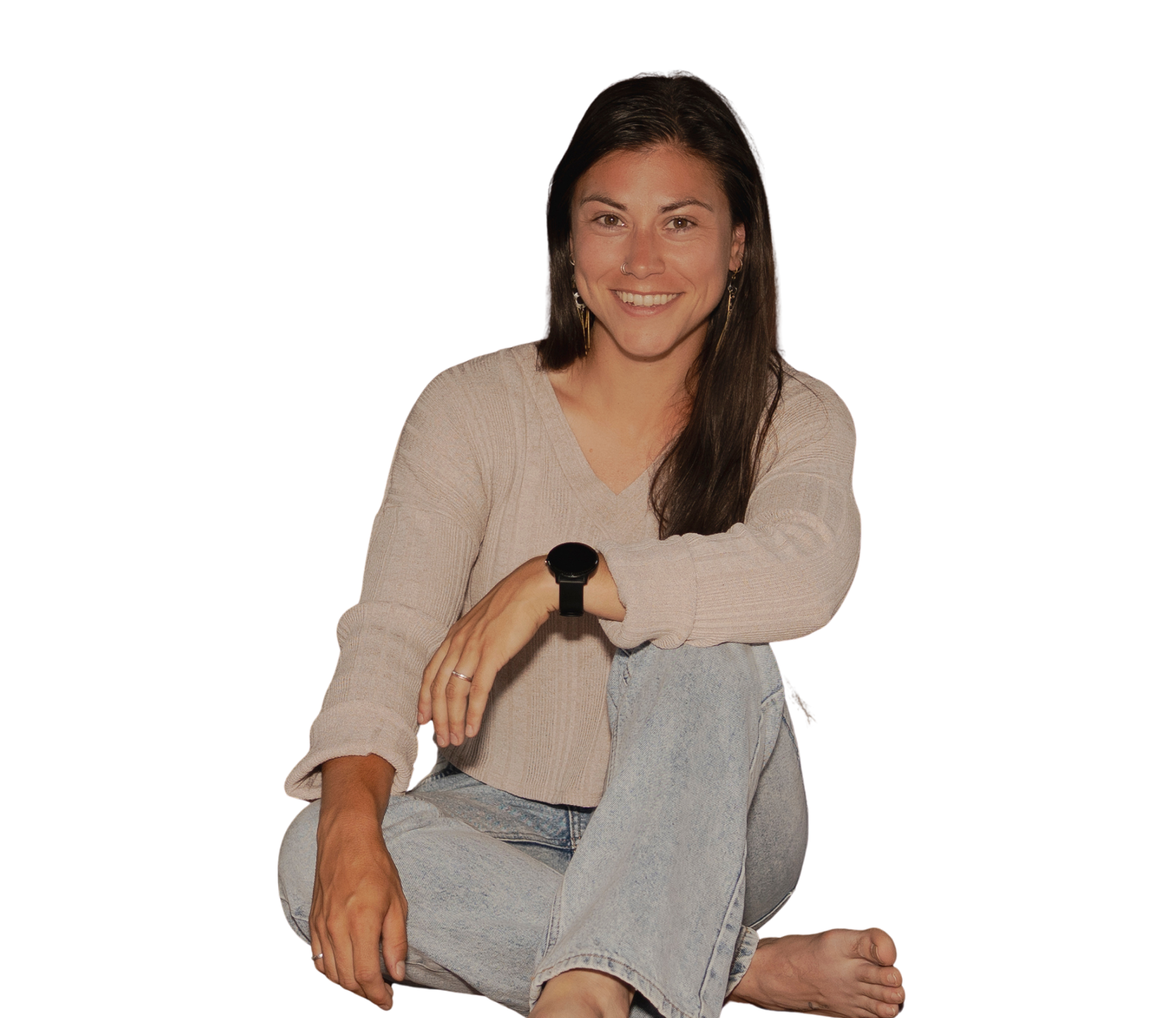 A smiling woman with dark hair, wearing a beige sweater and denim shorts, sitting with one leg crossed over the other and her arms resting on her knee, against a white background.