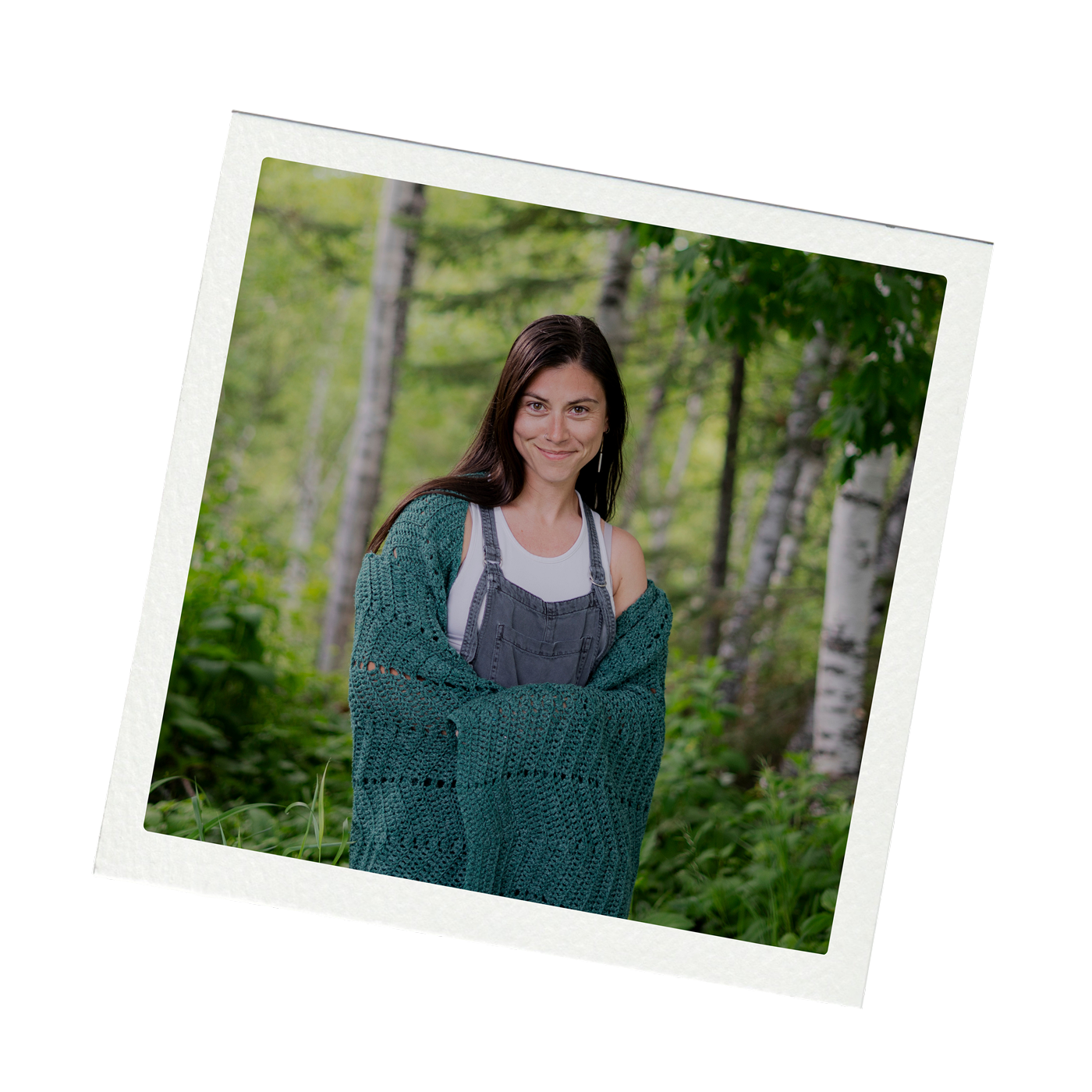A woman smiling in a forest setting with trees and greenery, wearing a white top, denim overalls, and a teal knitted cardigan.