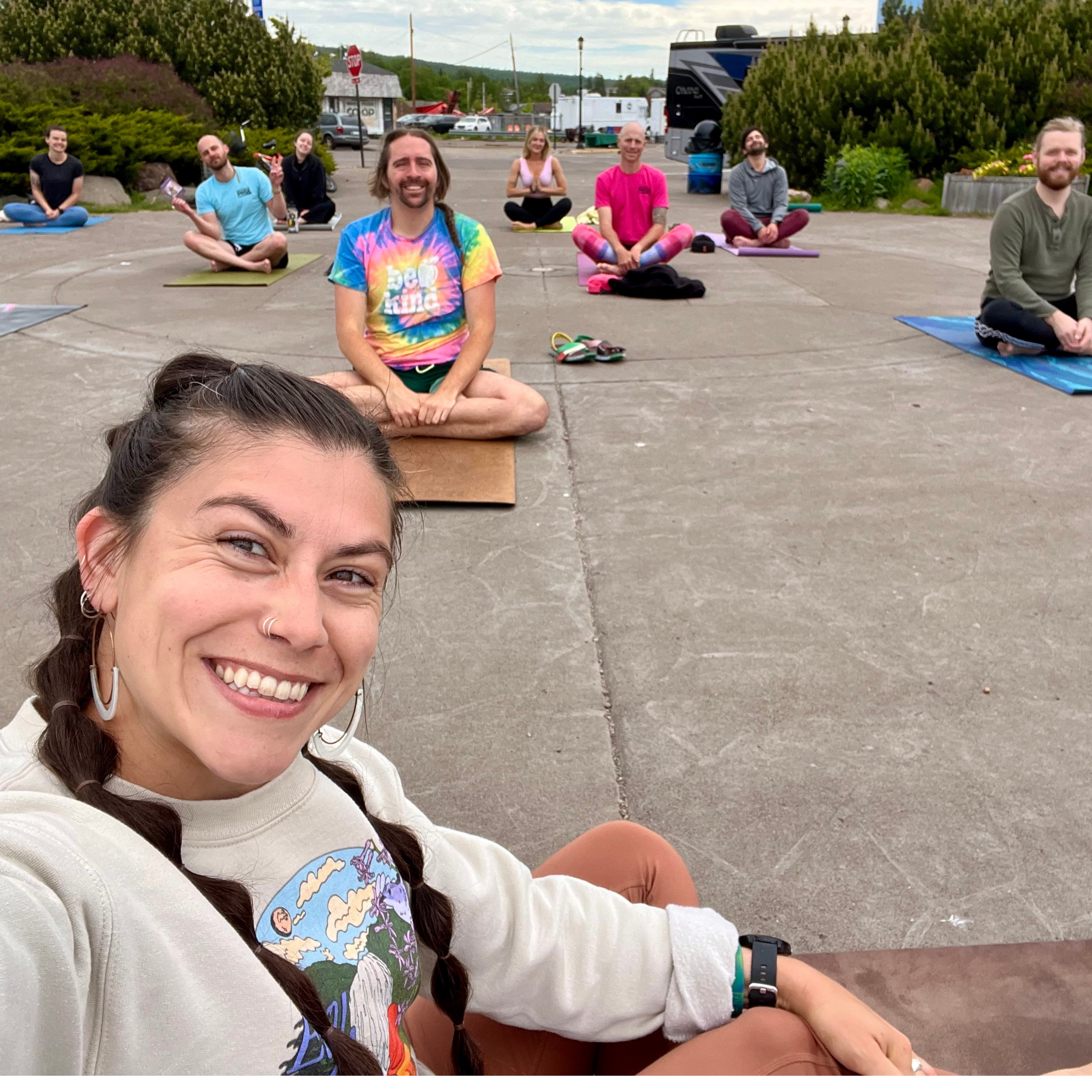 A group of people practicing yoga outdoors on a paved area with trees, bushes, and a stop sign in the background. The people are sitting cross-legged on yoga mats, with some smiling or meditating.