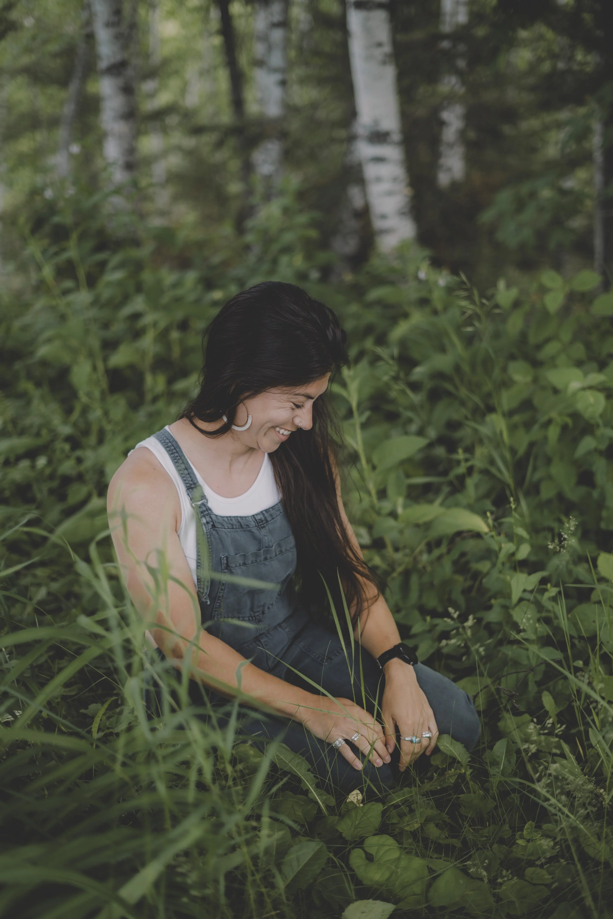 A woman sitting in a lush green outdoor setting with trees and foliage, smiling and looking down, wearing a white tank top and denim overalls.